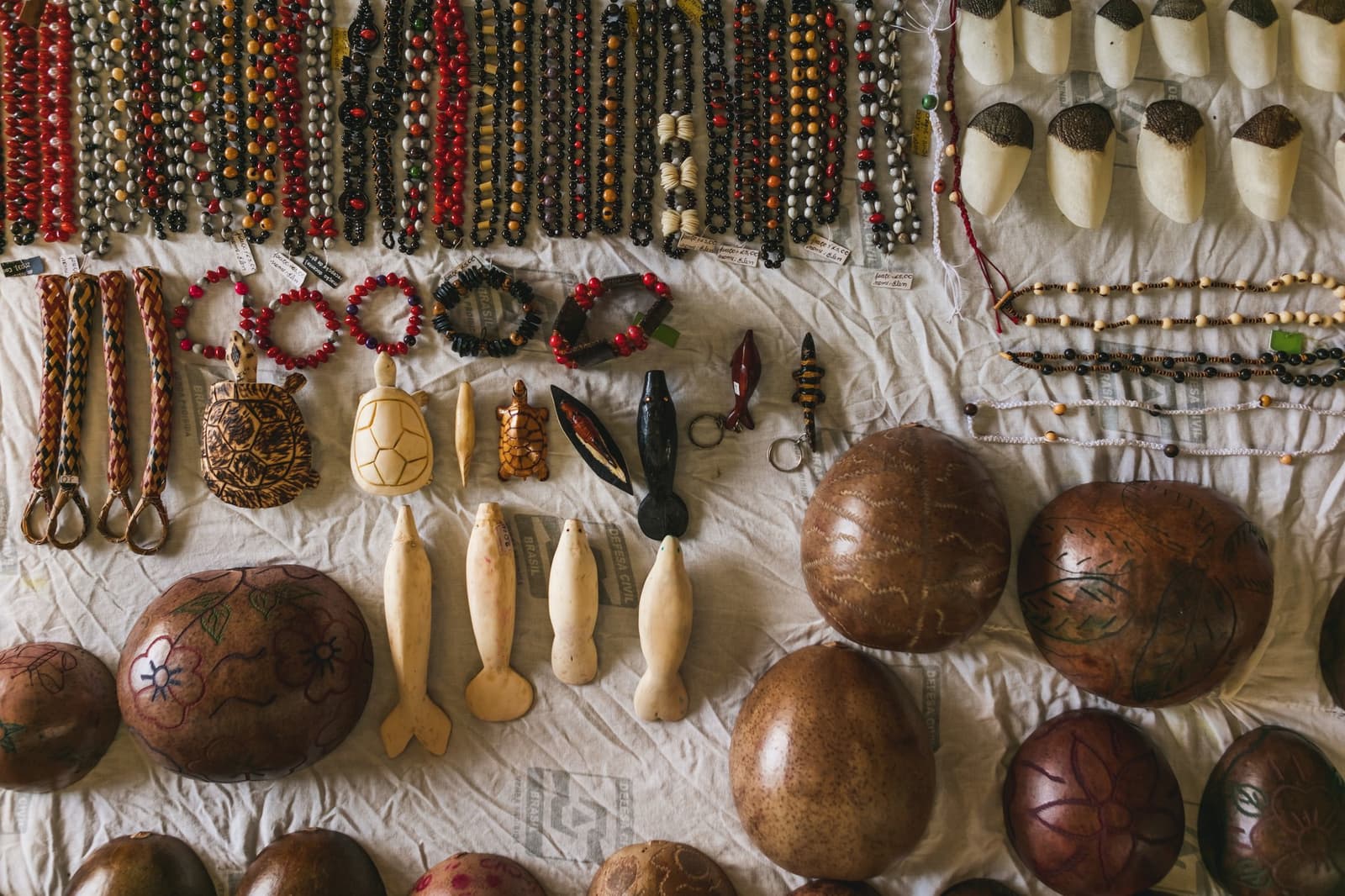 Met de hand gemaakt, met potlood geprijsd, neem rustig je tijd. Photo: Guilherme Gomes Bovenaanzicht van Amazoniaanse handgemaakte ambachten op een wit doek tijdens een amazon rainforest ecolodge reis in Brazilië, met zadenkralen halskettingen en armbanden in rood, zwart en oranje, gesneden schildpadschilden en dolfijnornamentjes, gevlochten sleutelhangers en beschilderde kalebasschalen