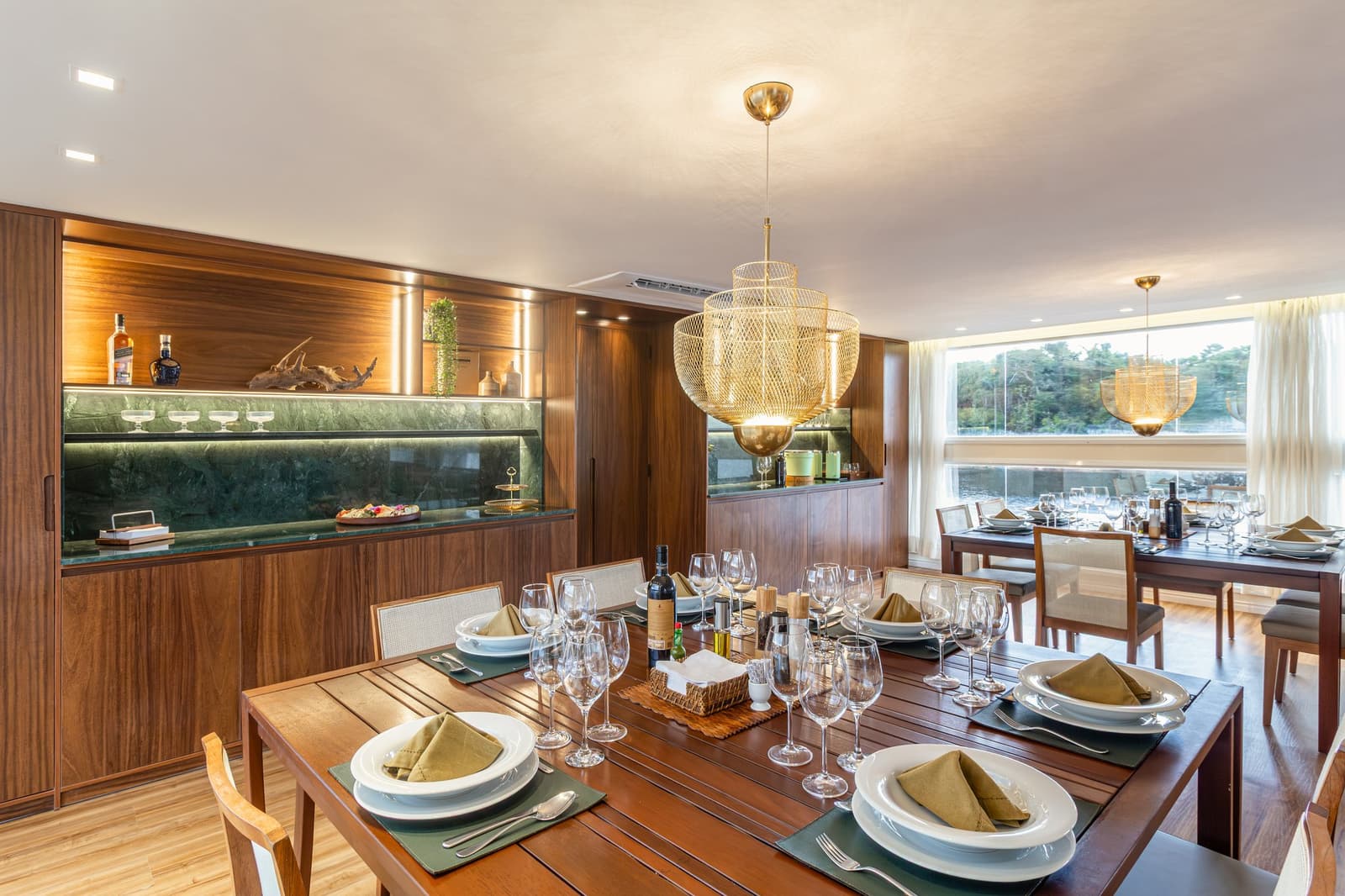 Lunchen met de Amazone als decor: dat maakt elk gerecht een beetje bijzonderder. Photo: STUDIO CREATE Elegant gedekte eetzaal aan boord van een luxe Amazone-cruise in Brazilië, met twee houten tafels vol witte borden, olijfgroene linnen servetten en wijnglazen, een verlichte walnoothouten bar met groen marmeren blad en grote ramen met uitzicht op de rivier