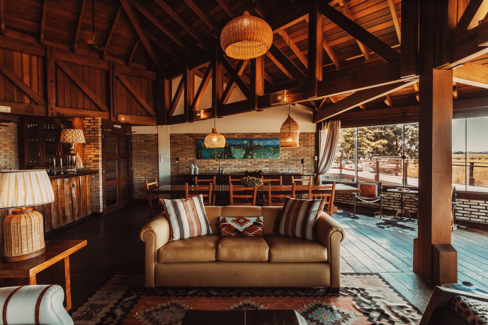 Nach dem Safari-Tag macht sich die Couch bezahlt. Photo: Felipe Castellari Offener Loungebereich einer Lodge auf einer Luxury Pantanal and Amazon Trip mit Gewölbedecke aus Holz, Korbpendelleuchten, einem beigefarbenen Ledersofa mit gemusterten Kissen, einem Kelim-Teppich und Panoramafenstern mit Blick auf Savanne und Holzterrasse