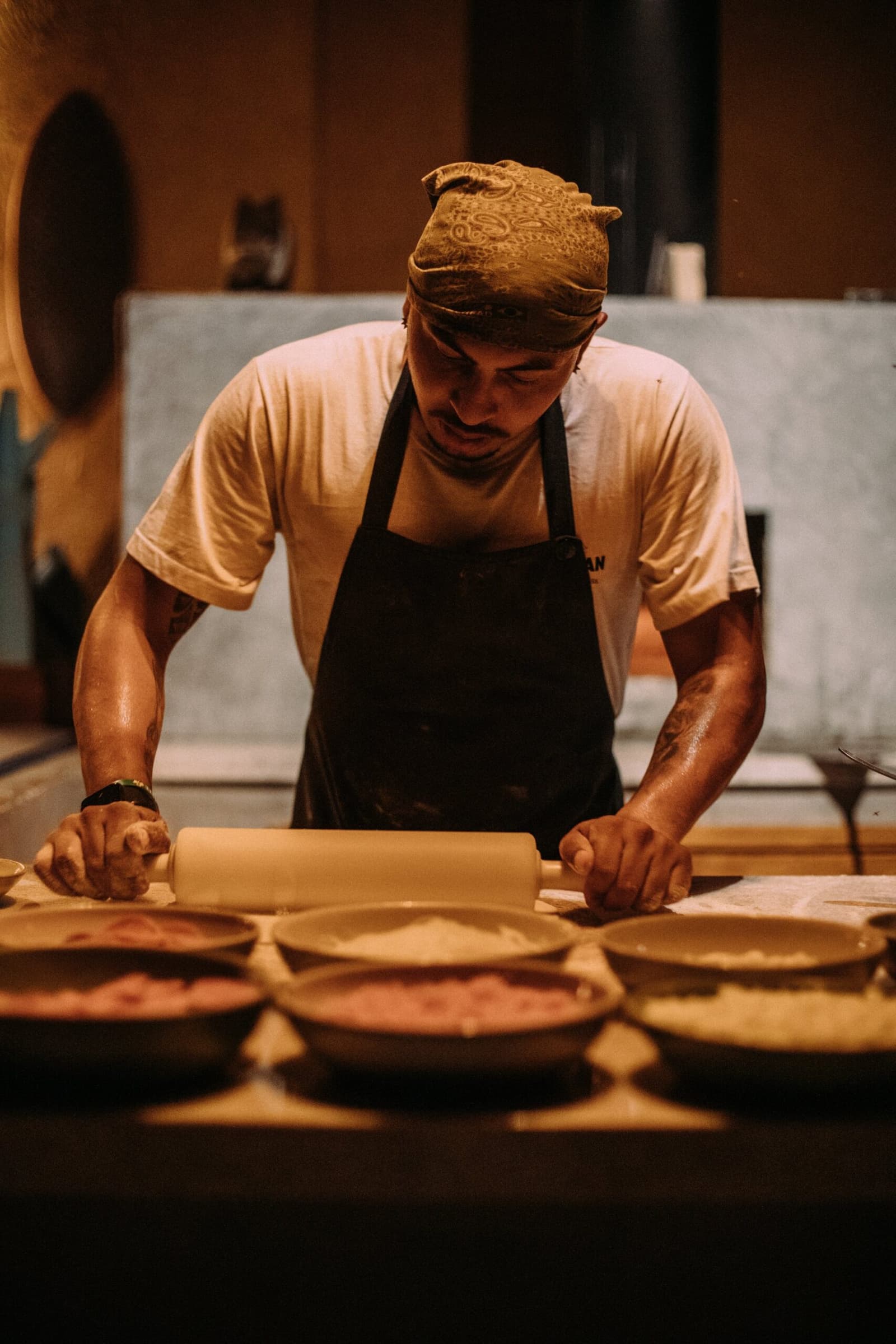 Tief im Teig, lange bevor der erste Gast am Tisch sitzt. Ein tätowierter Koch mit Bandana und dunkler Schürze rollt Teig auf einer Holztheke aus, während mehrere Keramikschüsseln mit Füllung vor ihm bereitstehen – Vorbereitung auf ein Lodge-Dinner