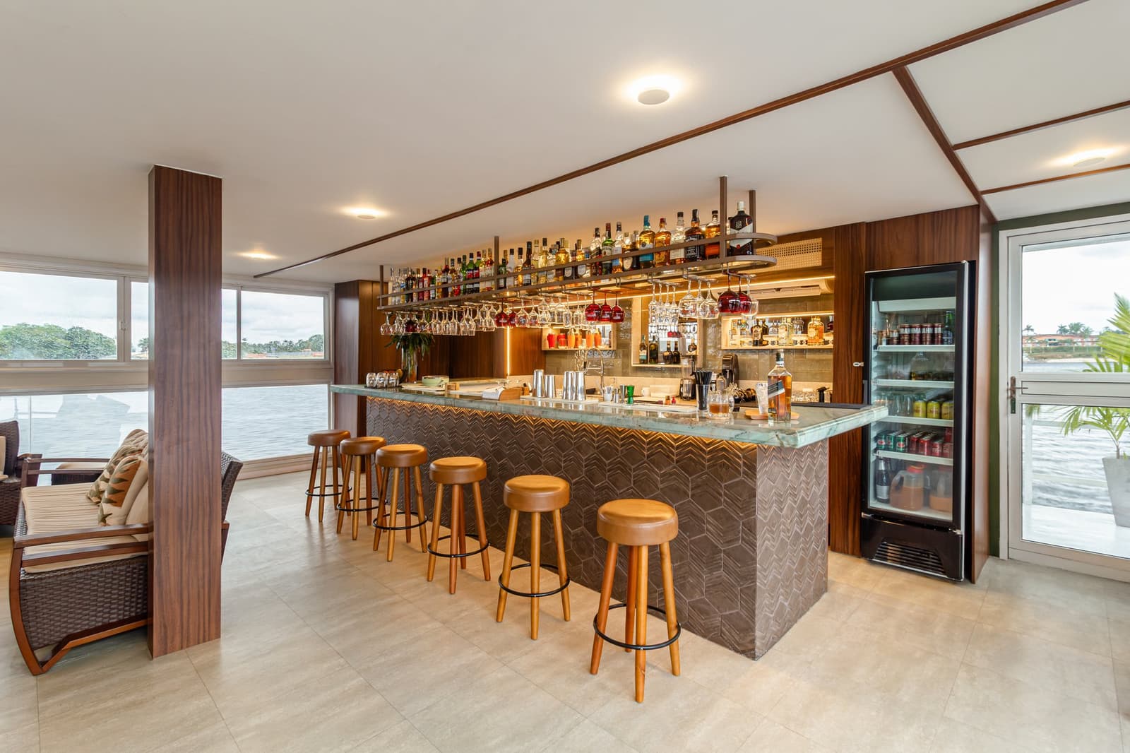 Na een dag op het water verdient zo'n bar zijn plek dubbel en dwars. Photo: STUDIO CREATE Goed gevulde bar aan boord van een luxe Amazone-cruise in Brazilië, met een groenmarmeren toonbank, verlichte schappen vol sterkedrank, hangende wijnglazen, leren barkrukken en panoramaramen met uitzicht op de rivier