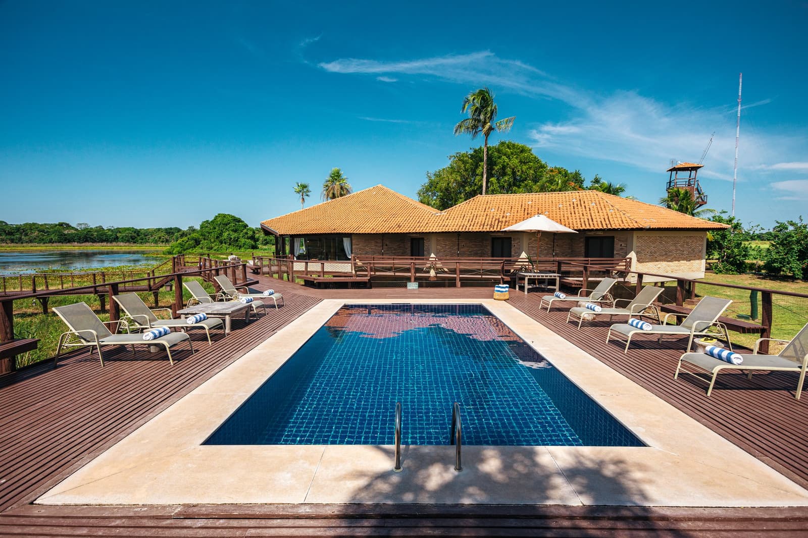 Zwischen zwei Safaris kurz abkühlen, der Pantanal hat seinen eigenen Rhythmus. Photo: Felipe Castellari Rechteckiger Pool auf einem Hartholzdeck mit Sonnenliegen, dem Hauptgebäude des Lodges und einem Blick auf einen Fluss im Pantanal im Hintergrund