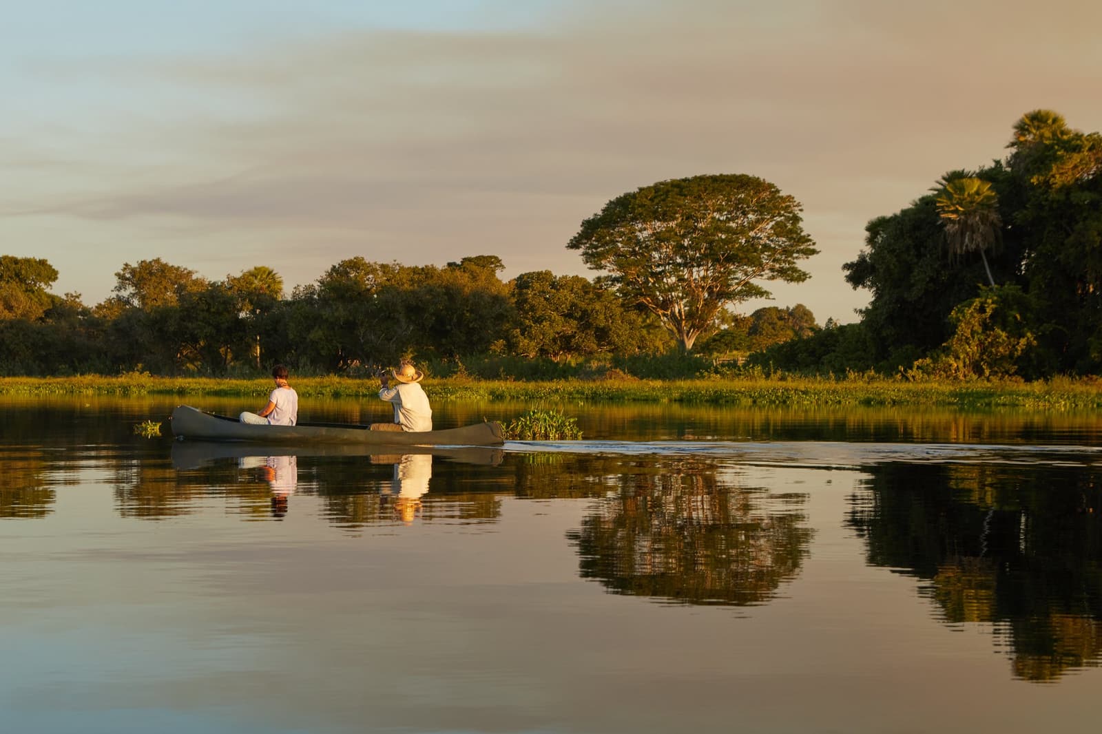 Im Abendlicht trägt der Fluss den Himmel auf seiner Oberfläche. Zwei Reisende sitzen ruhig in einem Kanu auf einem spiegelglatten Flusslauf im Feuchtgebiet während der goldenen Stunde, ihr Spiegelbild perfekt im stillen Wasser, üppiger Galeriewald am gegenüberliegenden Ufer