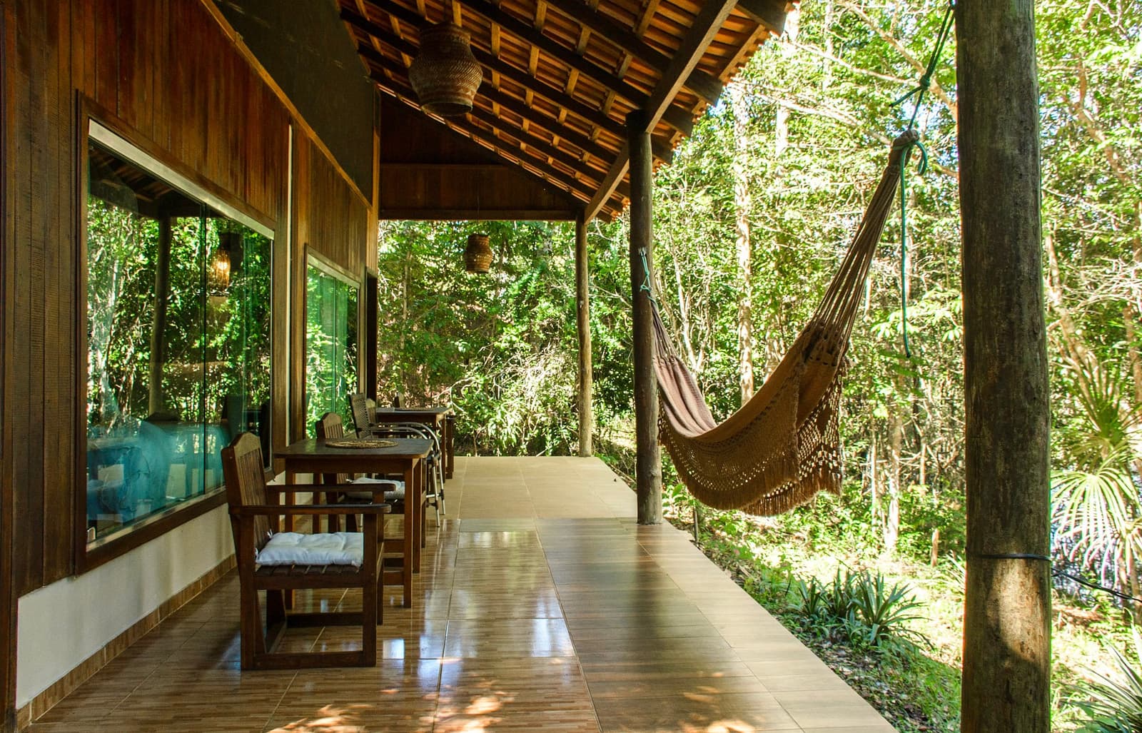 Een hangmat op ooghoogte met het bladerdak. Zo ziet een goede beslissing eruit. Overdekt terras van een Amazone-junglelodge met een katoenen hangmat met franje tussen houten palen, een teakhouten eettafel en stoelen, rieten hanglampen en dicht regenwoud vlak achter de open rand