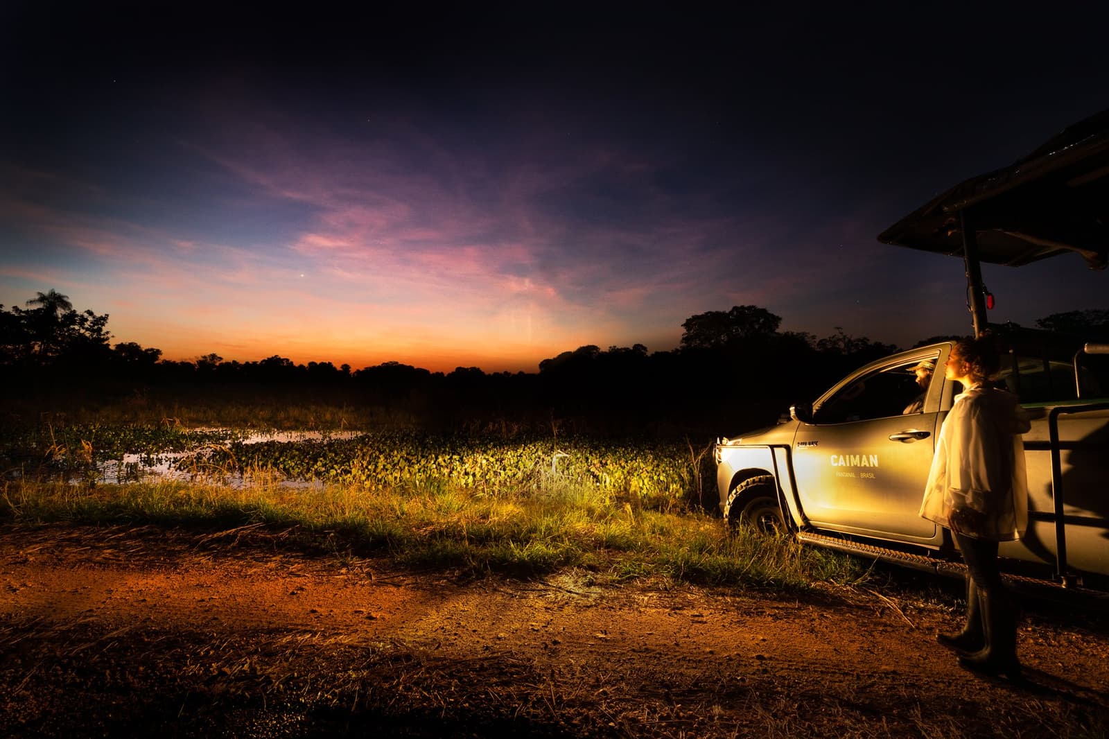 Genau in diesem Moment hört der Pantanal auf, still zu sein. Ein Guide steht neben einem Caiman-Lodge-Safariwagen auf einem Feldweg in der Abenddämmerung während einer Luxusreise durch den Pantanal und Amazonas, mit einem leuchtend orangefarbenen und violetten Abendhimmel über einem überfluteten Sumpf