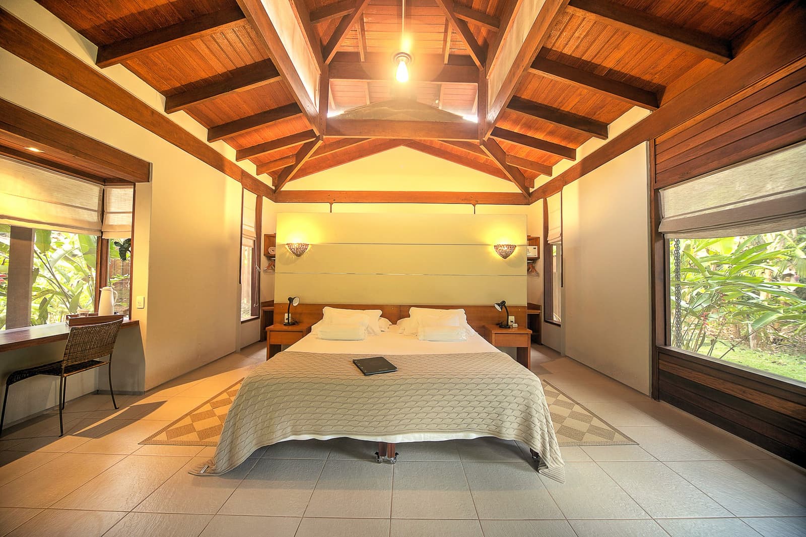 Hohe Decken, Grün direkt hinter der Scheibe, der Wald bleibt die ganze Nacht in Sichtweite. Photo: | SamuelMelim Photography Geräumiges Lodge-Zimmer mit gewölbter Holzbalkendecke, Doppelbett mit weißer Bettwäsche, warmen Wandleuchten und großen Fenstern mit Blick auf üppige tropische Vegetation