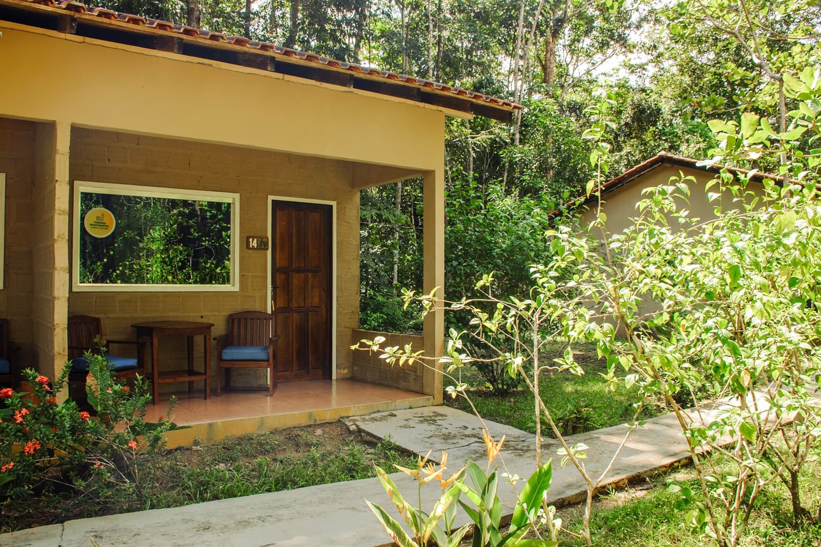 Kamer 14 heeft meer bomen dan buren, precies zoals het hoort. Buitenaanzicht van een genummerde junglebungalow met overdekt terras, houten stoelen en weelderig bos op de achtergrond, accommodatie op een Amazon- en Pantanal-tour