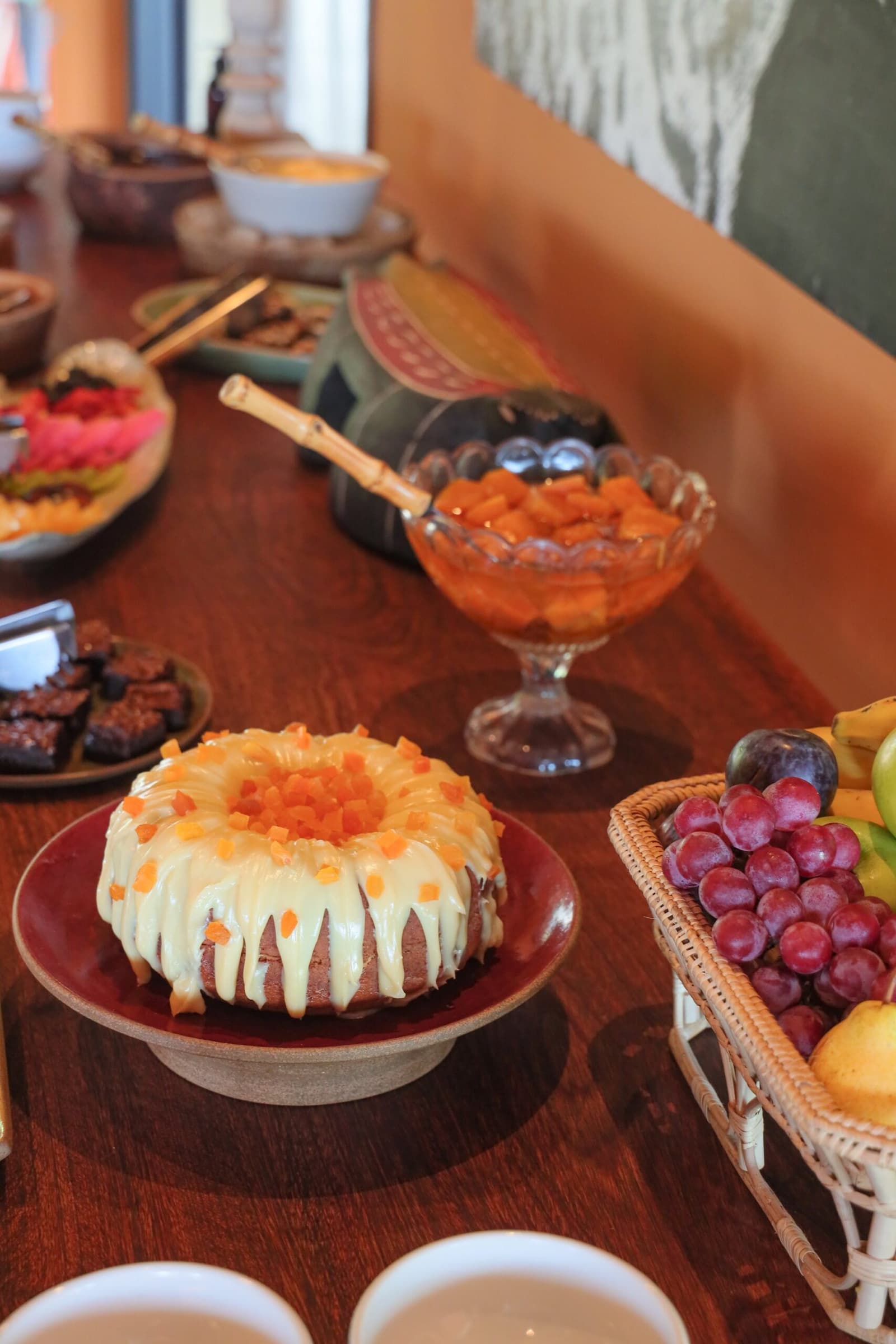 Erst Kuchen, dann Kaimane. Die Reihenfolge stimmt. Photo: thiagolbrito Ein glasierter Gugelhupf mit gewürfelter Papaya auf einem roten Keramikständer, umgeben von Obstschalen, Trauben, Brownies und einer Glasschale mit Orangenfrucht — üppiges Frühstücksbuffet auf einer Luxusreise durch Pantanal und Amazonas