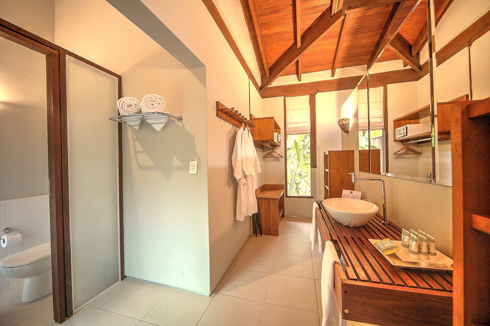 Knackig weiße Handtücher, Holzbalken, und draußen schon der Regenwald. Komfort, der nicht angeben muss. Photo: | SamuelMelim Photography Geräumiges Lodgebad mit freiliegenden Holzbalken an der Decke, Aufsatzwaschbecken auf einem Hartholz-Waschtisch, gefalteten weißen Handtüchern, Bademänteln und einem Fenster mit Blick ins Grüne