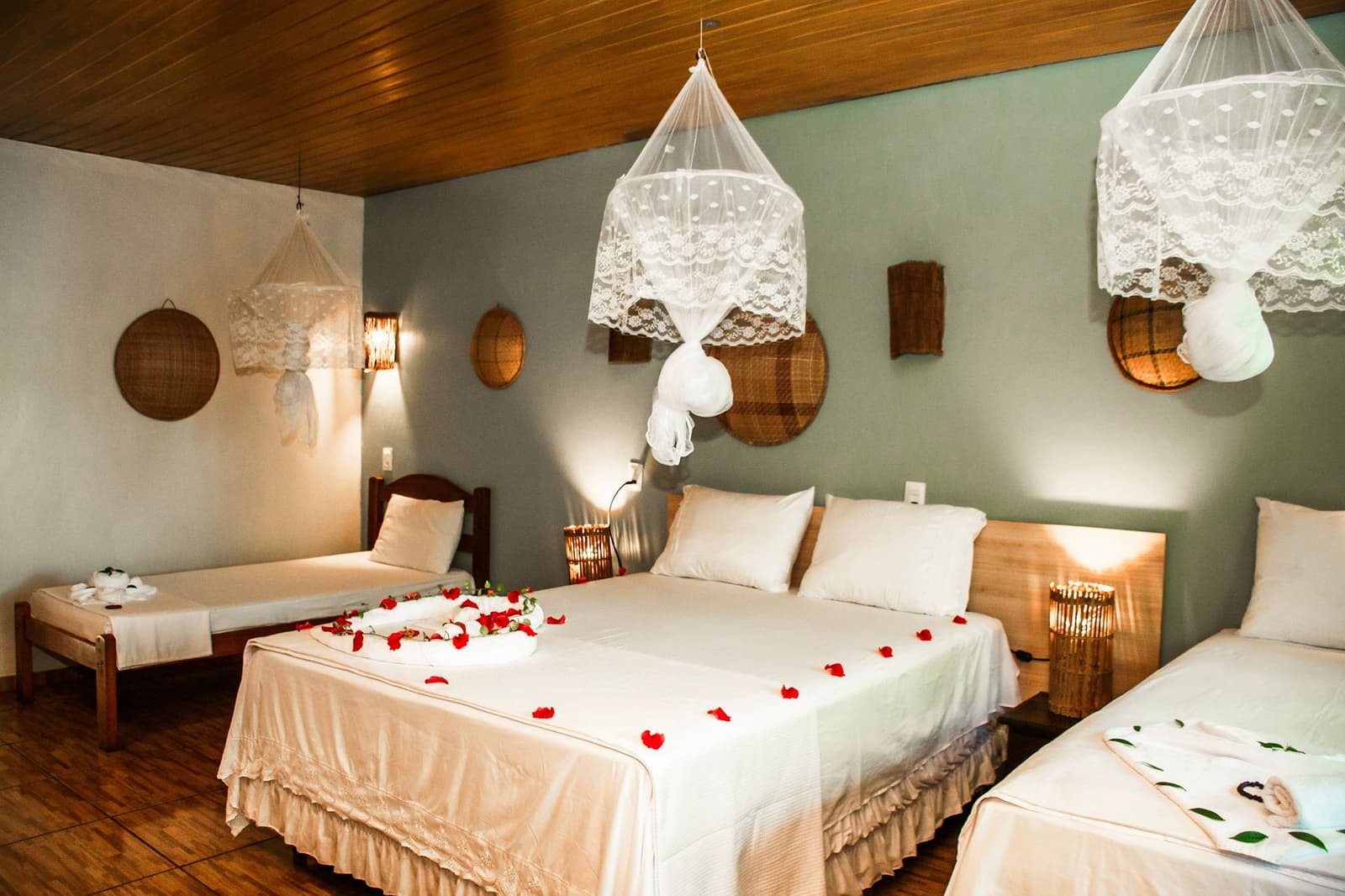 Rozenblaadjes, kanten klamboes en zacht licht. De ontvangst is duidelijk serieus genomen. Een kamer in een lodge voor een Amazon- en Pantanal-tour, met drie bedden met witte beddengoed bezaaid met rode rozenblaadjes, kanten klamboes aan een houten plafond, rieten wanddecoraties en warme bamboe lantaarns op de nachtkastjes.