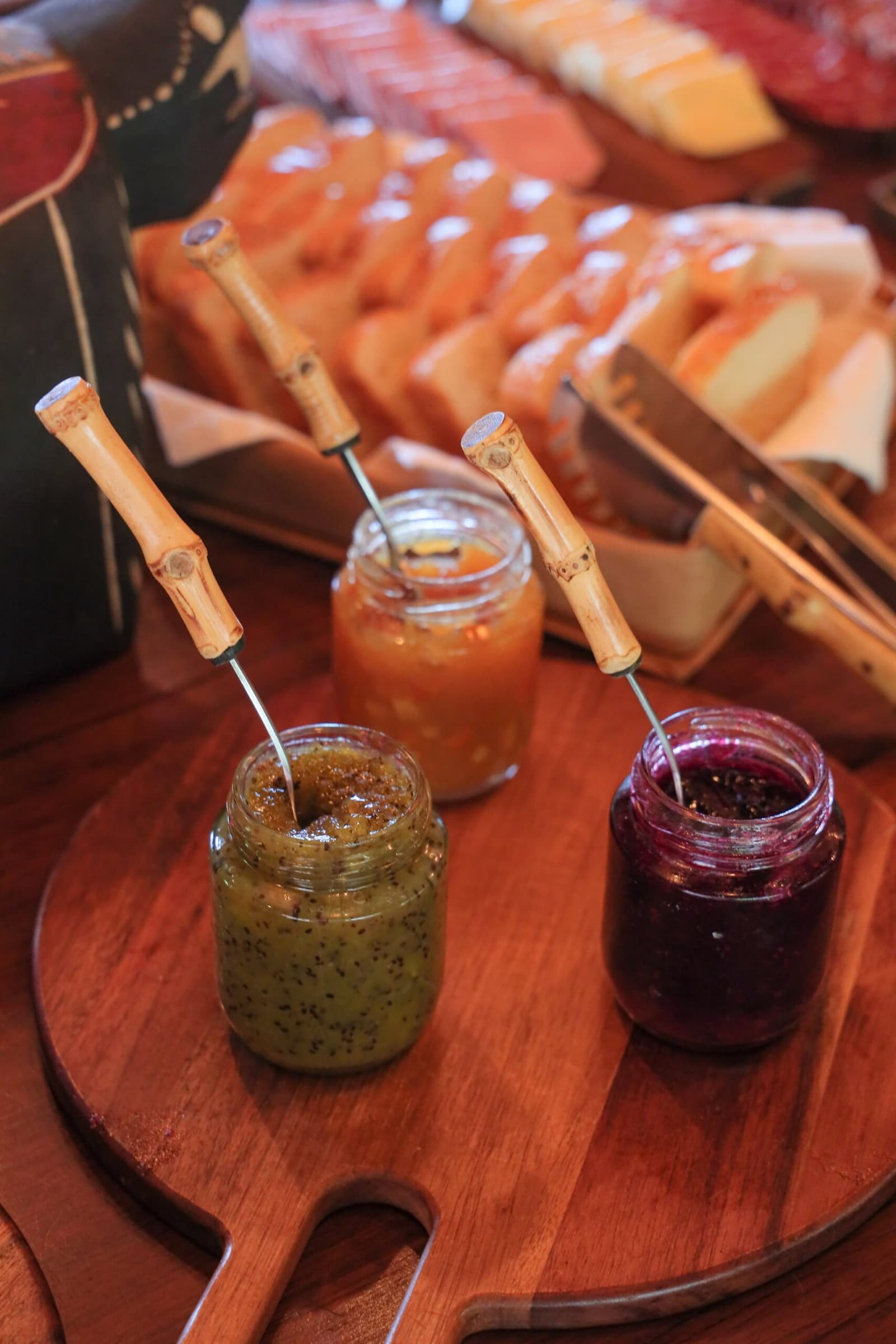 Drei Marmeladengläser, drei Aromen, und ein Brotkorb, der das Aufstehen verzögert. Photo: thiagolbrito Drei Glasgläser mit handwerklicher Fruchtmarmelade und Bambuslöffeln auf einem runden Holzbrett, mit Brotscheiben, Papaya und Käse im Hintergrund auf einem Lodge-Frühstückstisch