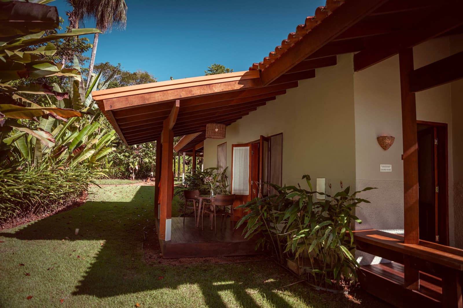 Schatten, ein gedeckter Tisch und der Dschungel beginnt direkt hinter dem Rasen. Ein Bungalow mit Ziegeldach auf einem Luxus-Pantanal-und-Amazonas-Trip, mit überdachter Holzveranda, geflochtenem Pendelleuchte, Esstisch im Freien und üppigem tropischem Garten unter blauem Himmel