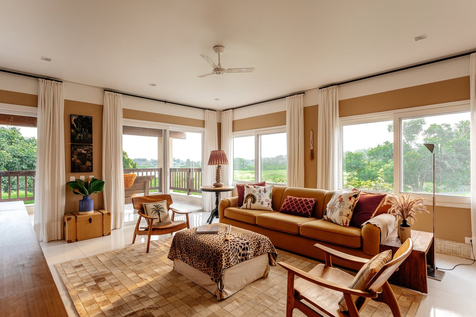 Warme Erdtöne, weiter Blick ins Grüne, und ein Deckenventilator, der seinen Job kennt. Photo: Felipe Castellari Helles Lodge-Wohnzimmer auf einer Luxusreise durch Pantanal und Amazonas mit karamellfarbenem Sofa, bedruckten Kissen, einem Leopardenmuster-Hocker, Holzsesseln und raumhohen Fenstern mit Blick auf Feuchtgebiete und grüne Baumkronen