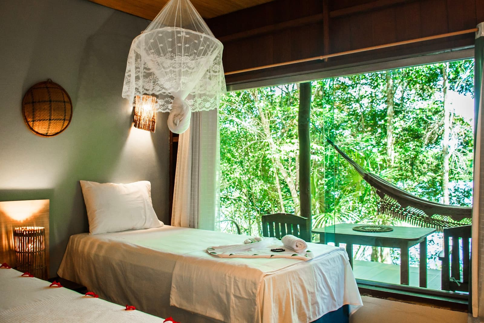 Kanten netten, fris linnengoed en een hangmat die buiten op je wacht. Tweepersoonskamer in een Amazone junglelodge met witte linnen bedden onder een kanten muskietennet, warme wandlamp, gevlochten mandendecoratie en een groot raam met uitzicht op een groen bladerdak en een hangmat op het balkon