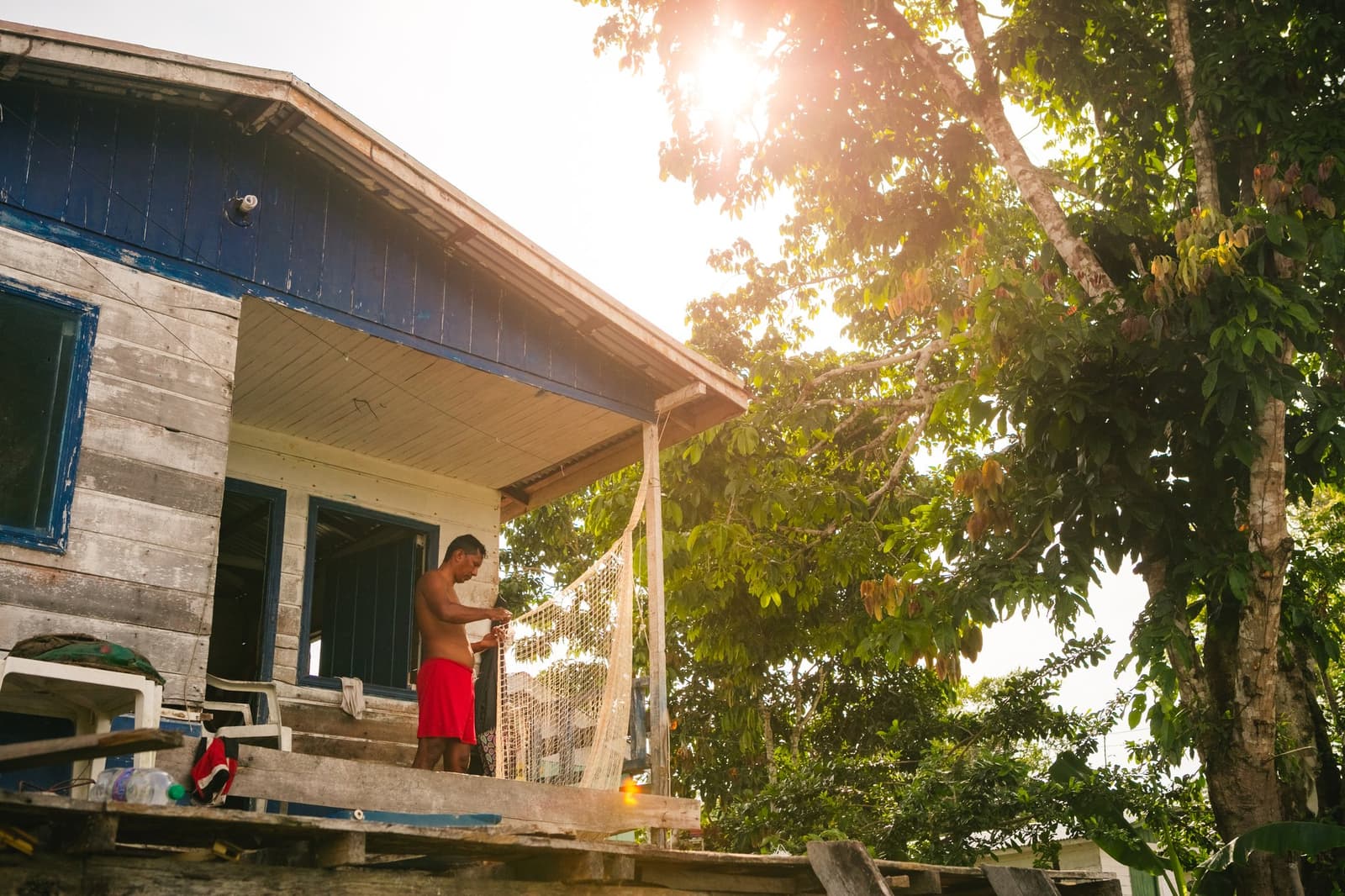 Netten lappen zichzelf niet op, en het ritme hier wacht niet op toeristen. Photo: Guilherme Gomes Een man herstelt een visnet op de veranda van een verweerd blauw-grijs houten huis naast het bos, met warm tegenlicht dat door het bladerdak filtert, tijdens een amazon rainforest ecolodge trip in Brazilië met een bezoek aan een riviergemeenschap