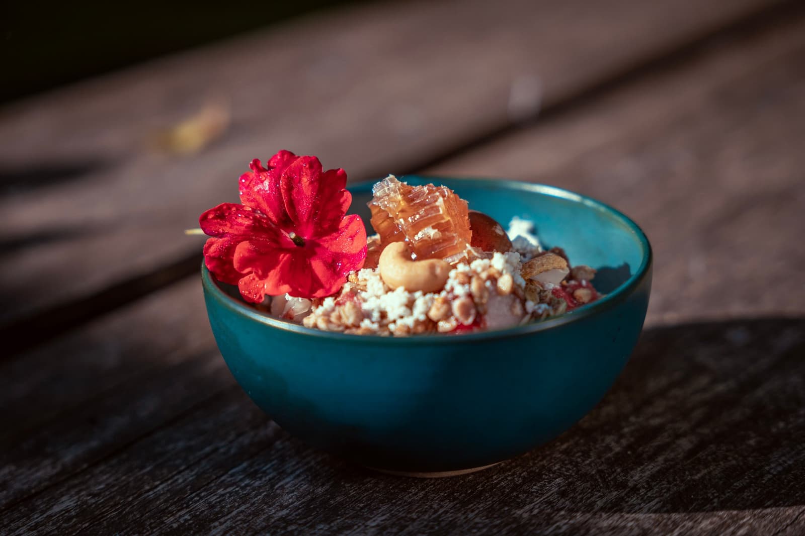 Frühstück, das sich Zeit lässt, Honigwabe, Nüsse und eine Blüte, einfach weil es geht. Eine blaugrüne Keramikschale auf einem verwitterten Holztisch, gefüllt mit Granola, Nüssen, zerbröckeltem Weißkäse und einem Stück roher Honigwabe, garniert mit einer leuchtend roten essbaren Blüte
