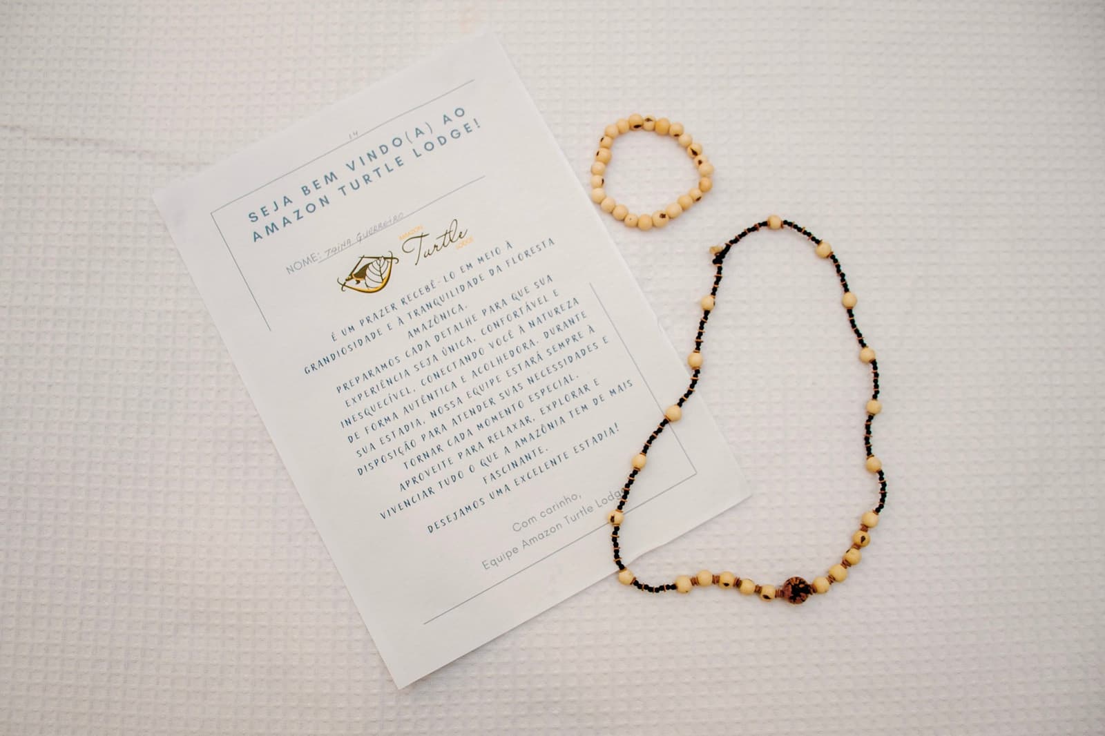 Zaden met de hand geregen, een kaartje met jouw naam. Ze wisten dat je kwam. Gepersonaliseerde welkomstkaart van een Amazone-lodge naast een handgemaakte zaadketting en bijpassende armband op een wit wafelgeweven beddengoed