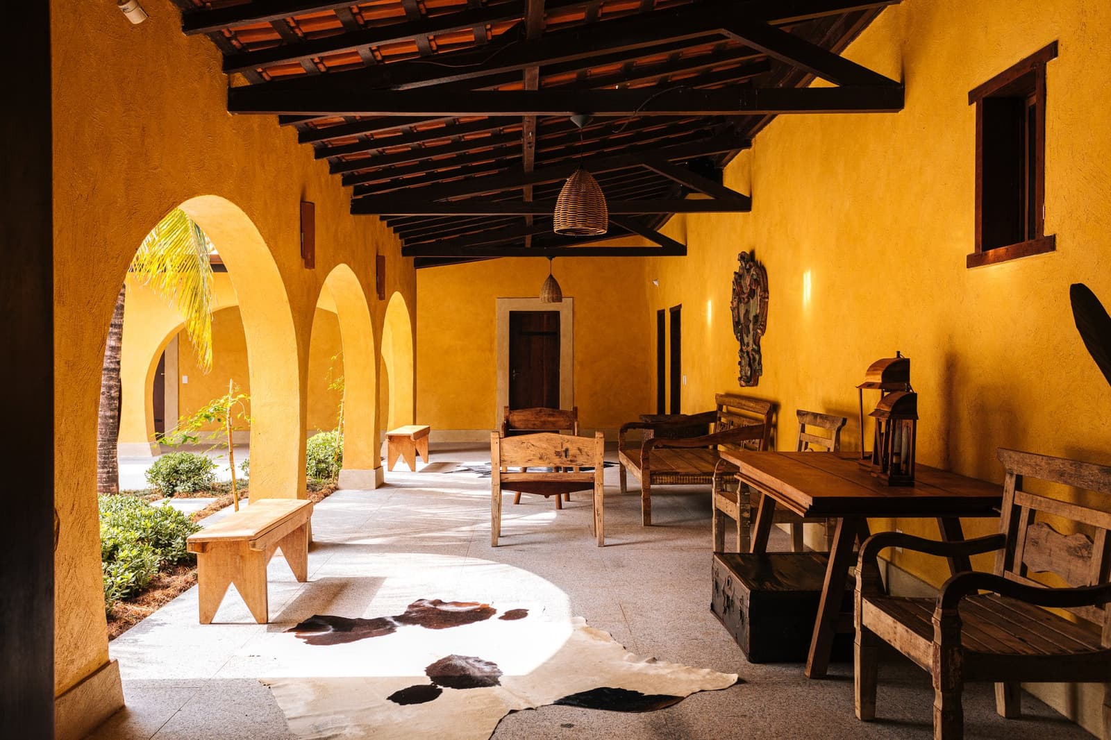 Bögen, Holzbalken und ein Kuhfell, Komfort mit ganz eigenem Charakter. Überdachte Veranda einer Lodge für Luxus-Pantanal-und-Amazonas-Reisen, mit goldgelben Bogenarkaden, dunklen Holzbalken, rustikalen Hartholzmöbeln und einem Kuhfellläufer auf dem Steinboden