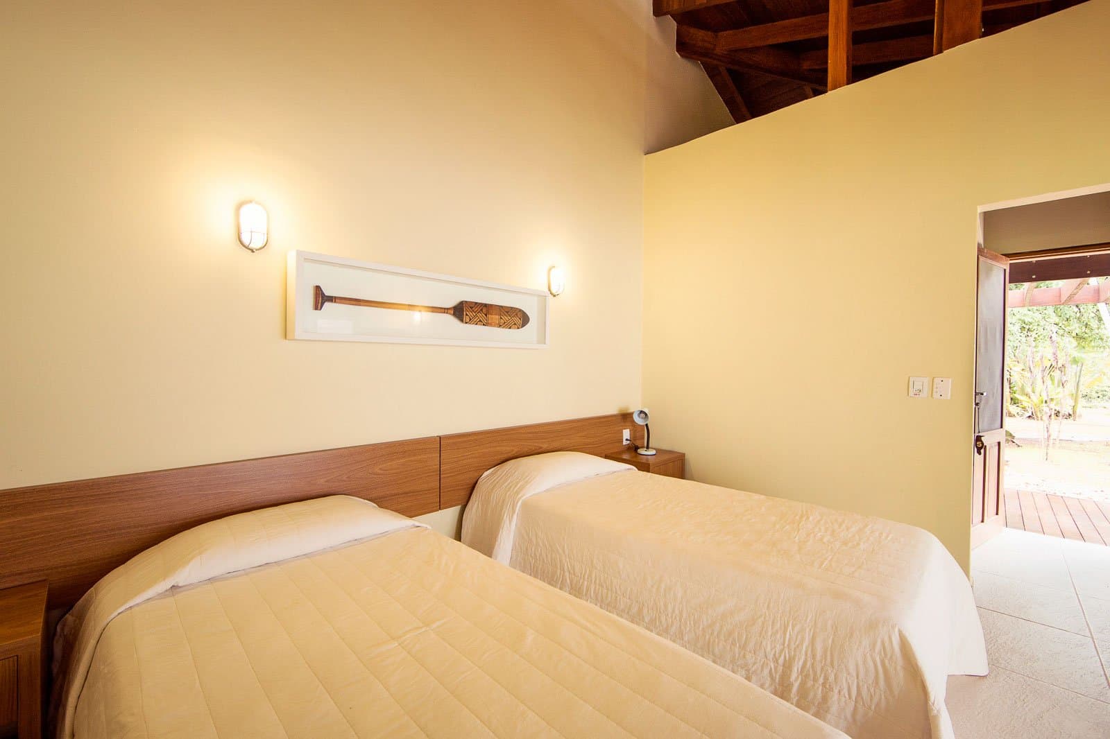 Zwei Betten, ein Paddel an der Wand, und draußen wartet der Wald. Photo: | SamuelMelim Photography Zweibettzimmer in einer Lodge für eine Luxus-Reise in Pantanal und Amazonas, mit cremefarbenen Steppdecken, warmem Holzkopfteil, Wandleuchten, einem gerahmten dekorativen Paddel an der Wand, freiliegenden Holzbalken an der Decke und einer offenen Tür zu einer sonnigen Terrasse