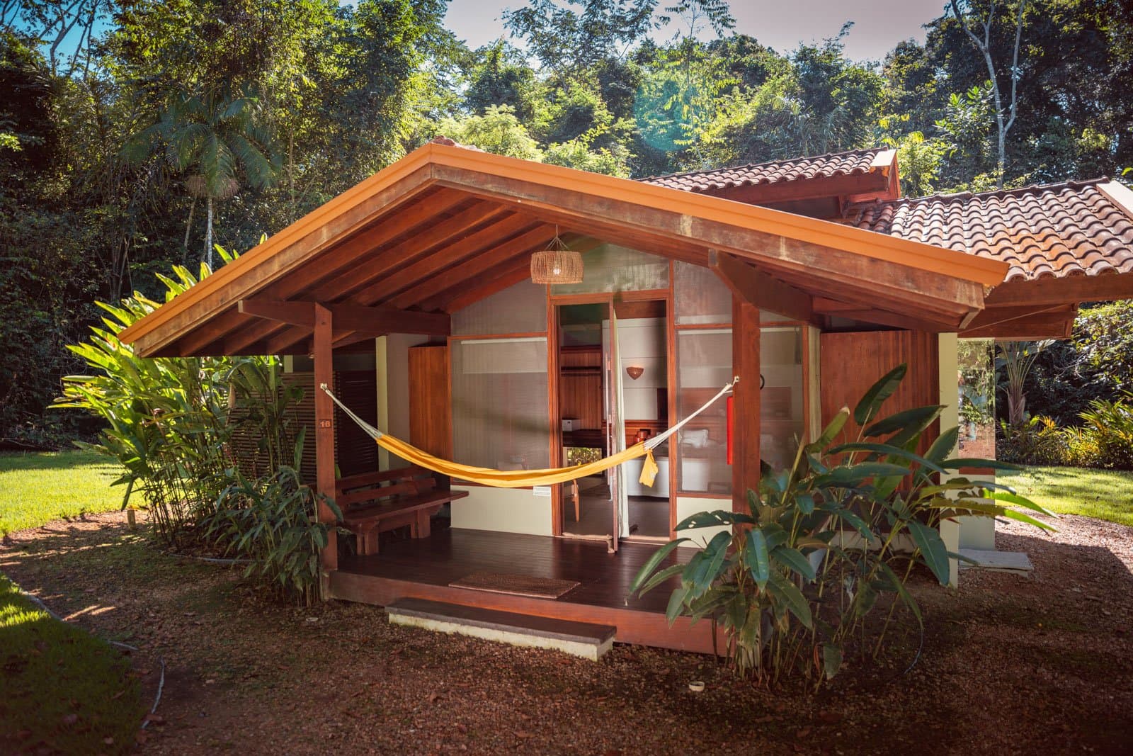 Eine gelbe Hängematte, eine Holzterrasse, und der Wald gleich nebenan. Photo: Samuel Melim Ein holzgerahmter Ökolodge-Bungalow mit gelber Hängematte auf der überdachten Veranda und dichtem tropischen Regenwald im Hintergrund