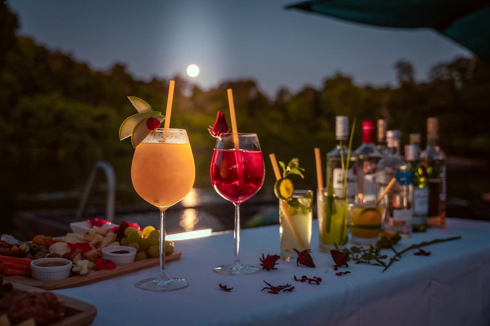 Vollmond, vier Cocktails, eine Käseplatte. Der Abend hat sich selbst organisiert. Vier garnierte Cocktails und ein Snack-Brett auf einem weißgedeckten Tisch in der Dämmerung, mit einem Vollmond über den Baumwipfeln auf einer Luxusreise durch Pantanal und Amazonas