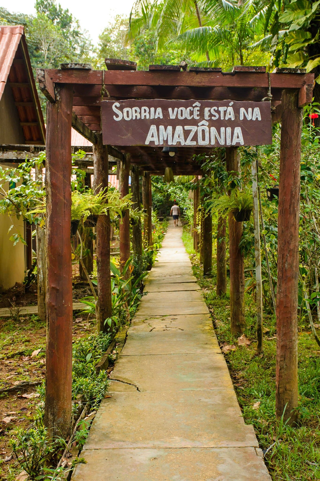 Het bos verwelkomt je op zijn eigen manier. Glimlach maar. Een rustieke houten pergola bij de ingang van een Amazone-junglelodge toont een bord met de tekst 'Sorria você está na Amazônia' — Lach, je bent in het Amazonegebied — met een met bomen omzoomd betonnen pad dat het regenwoud in leidt.