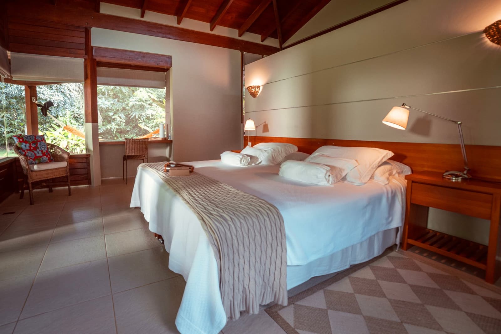 Weiche Kissen, warmes Holz, und der Wald drückt fast gegen die Scheibe. Geräumiges Lodgezimmer auf einer Luxusreise durch Pantanal und Amazonas, mit Kingsize-Bett in weißer Bettwäsche, warmen Holzdeckenbalken, Nachttischlampen und großen Fenstern zum dichten Grün des Waldes