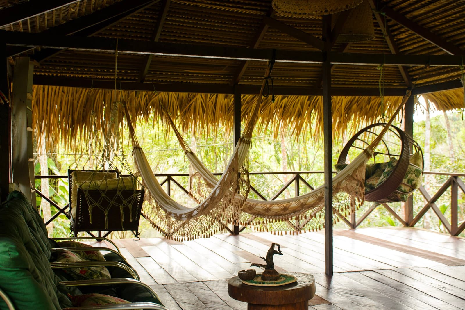 Kies een hangmat, welke dan ook. Moeilijker dan het klinkt. Open terras van een junglelodge met meerdere gefranjerde hangmatten, een rieten hangstoel en een palmdakbedekking met uitzicht op weelderig bos
