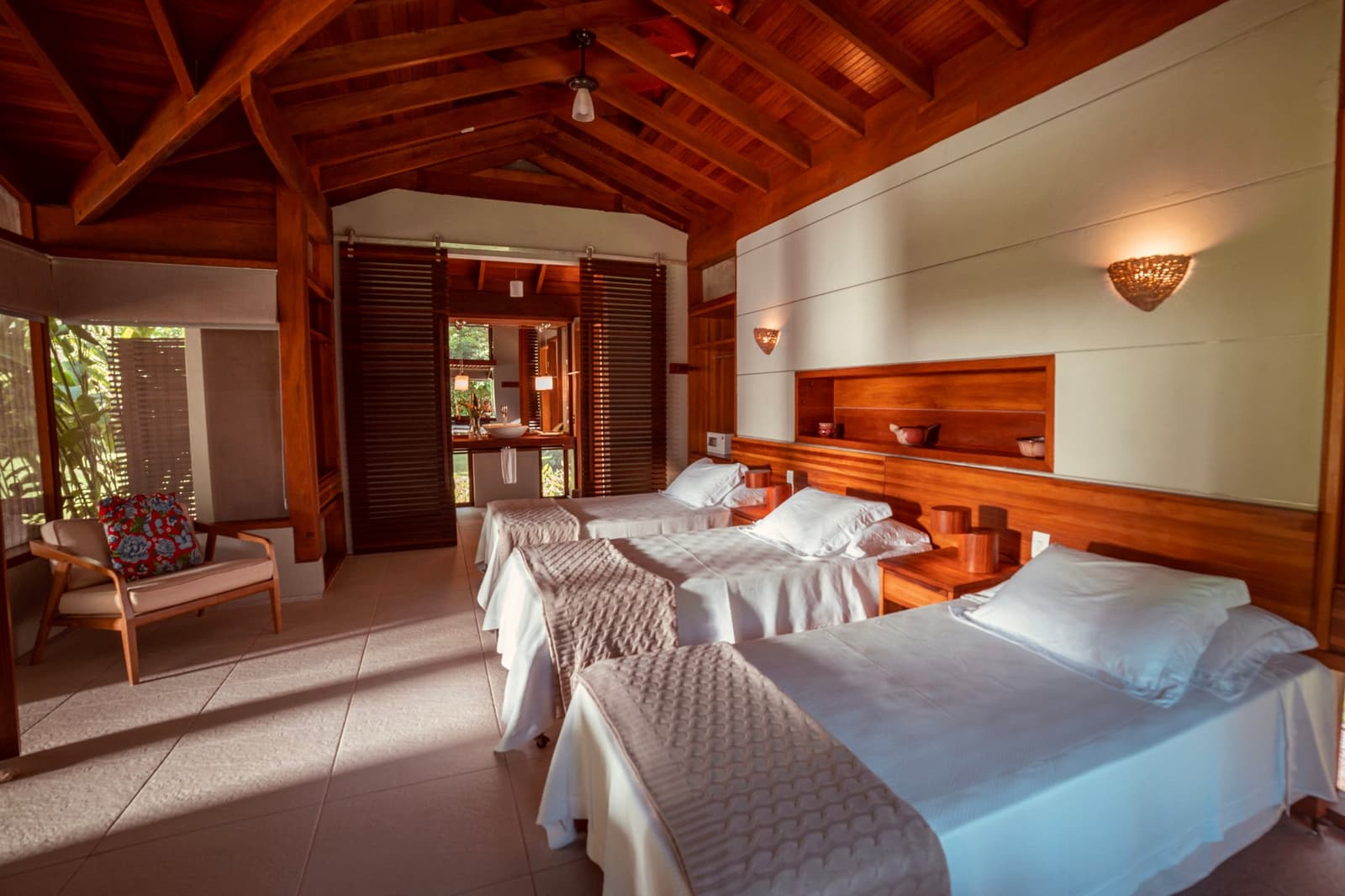 Holz, Ruhe und ein Deckenfächer, der genau das Richtige tut. Photo: Samuel Melim Zweibettzimmer einer Luxuslodge mit hohem Holzbalkengewölbe, warmem Holzkopfteil mit eingebauten Regalen, geflochtenen Wandleuchten und einer Schiebetür mit Lamellen, die zu einem En-suite-Bad führt, mit tropischem Gartensicht durch die Fensterläden