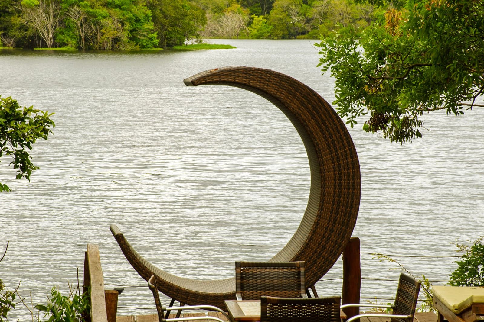 Die stoel is ontworpen door iemand die de oever nooit wilde verlaten. Een maanvormige rieten loungestoel op een houten terras met uitzicht op een rustige rivier omzoomd door dicht groen woud, bij een lodge van een Amazon en Pantanal tour