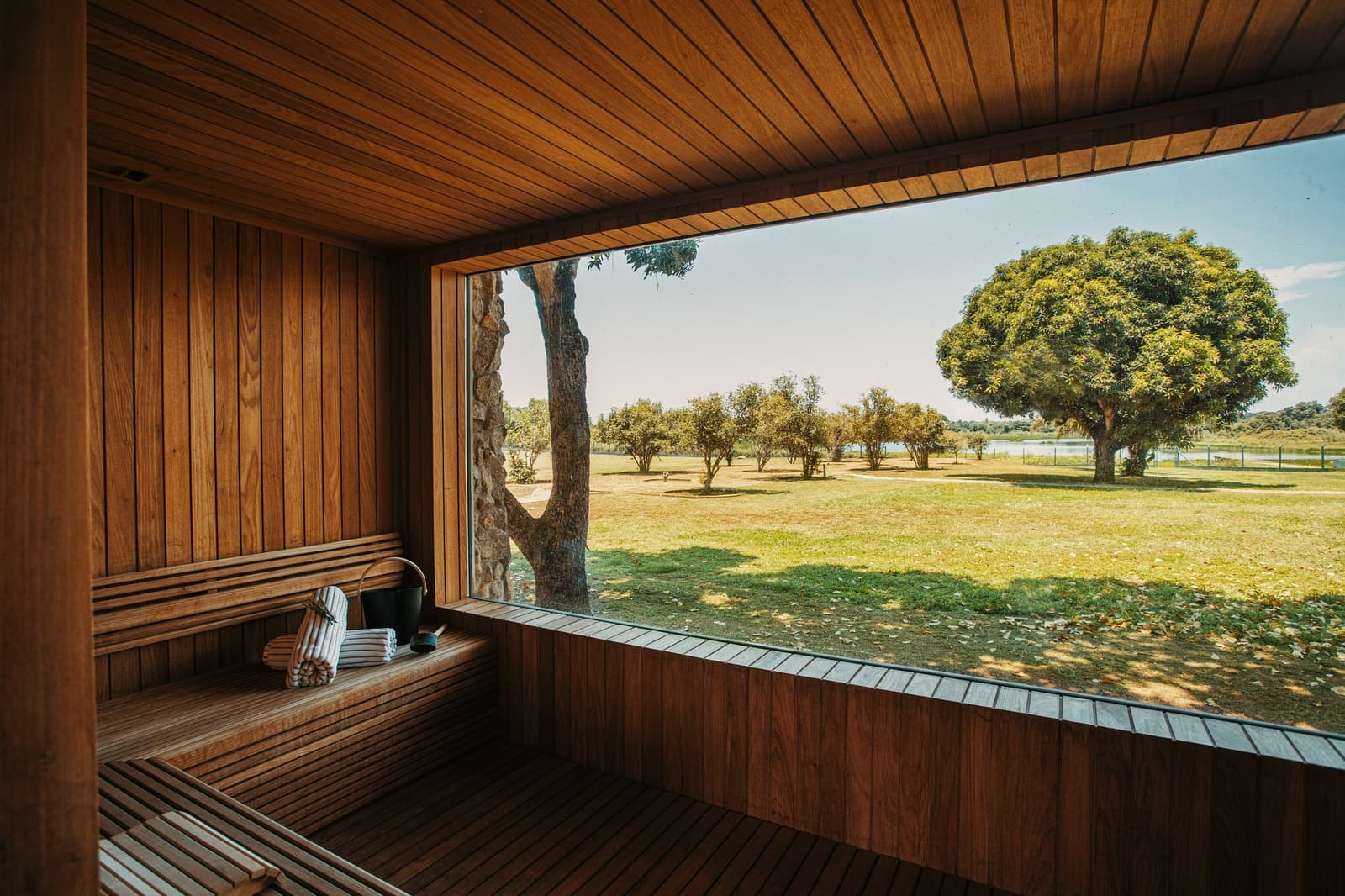 Schwitzen mit Aussicht, das Savannenpanorama gehört inklusive. Holzverkleidete Sauna-Innenraum auf einer Luxury Pantanal and Amazon Trip mit Panoramafenster und Blick auf sonnenbeschienene Graslandschaft, vereinzelte Bäume und eine entfernte Lagune