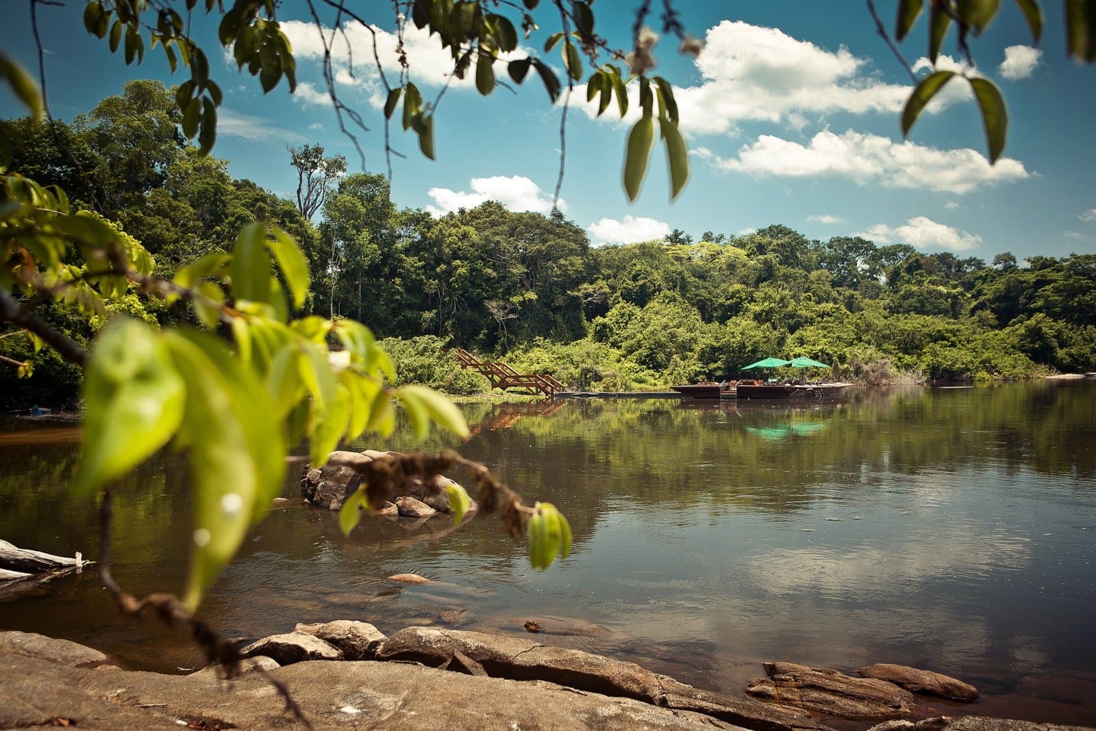 Felsen, Fluss und ein schattiger Steg, der sich nach einer echten Entdeckung anfühlt. Photo: Samuel Melim (c) Ein ruhiger Fluss im Regenwald spiegelt grünes Blätterdach und blauen Himmel wider, während eine hölzerne Uferlodge-Plattform mit türkisfarbenen Sonnenschirmen durch überhängende Blätter sichtbar ist.