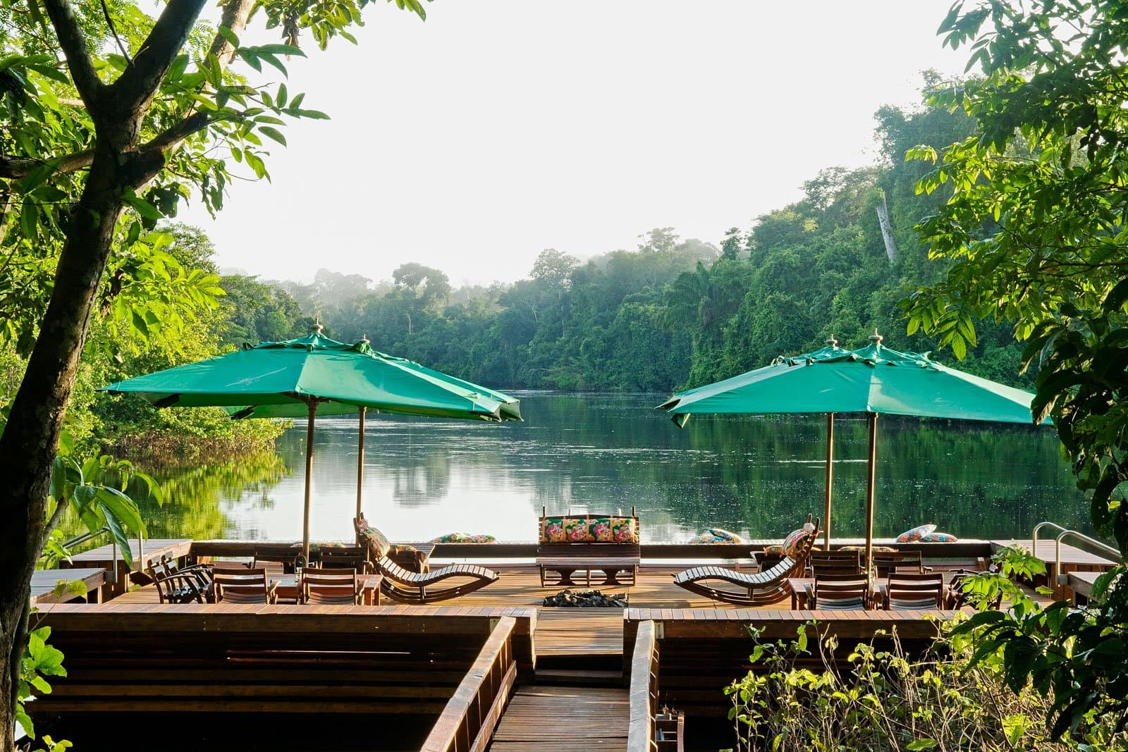 Der Wald reicht bis ans Ufer, den Rest erledigt der Fluss. Photo: Luis Gomes Eine Holzterrasse mit Liegestühlen und grünen Sonnenschirmen blickt auf einen ruhigen Fluss, der von dichtem Regenwald gesäumt wird — Teil einer Luxusreise durch den Pantanal und den Amazonas