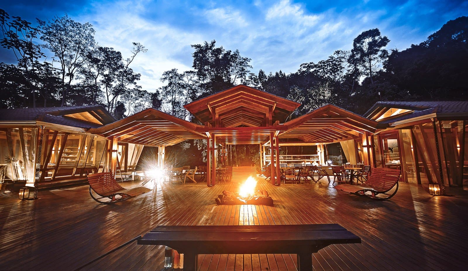 Ein Feuer, ein Drink, und der Wald bestimmt, wann der Abend endet. Photo: Samuel Melim (c) Offene Holzterrasse einer Lodge in der Abenddämmerung auf einer Luxusreise durch Pantanal und Amazonas, mit einem Lagerfeuer in der Mitte, Schaukelstühlen, einer beleuchteten Bar und Essbereichen unter spitzen Holzdächern, umgeben von hohen Bäumen vor blauem Zwielichtshimmel
