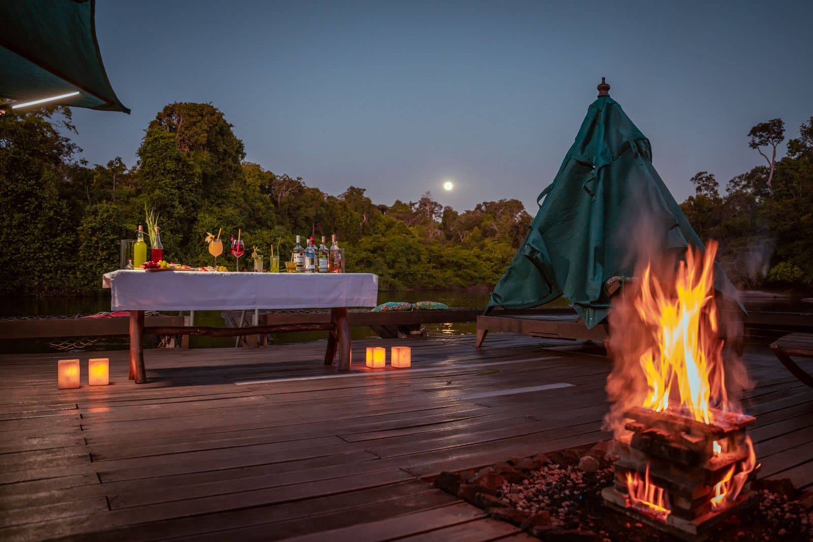 Cocktails, Kerzenschein, Vollmond, der Wald braucht keine große Bühne. Photo: Samuel Melim Eine Holzterrasse an einem Waldfluss in der Abenddämmerung, eingerichtet für einen Abend auf einer Luxusreise durch Pantanal und Amazonas: im Vordergrund ein prasselndes Lagerfeuer, Papierlaterne-Kerzen entlang der Dielen und ein mit weißem Leinen gedeckter Tisch voller Cocktails und Spirituosen, während ein Vollmond über den Baumkronen aufgeht.