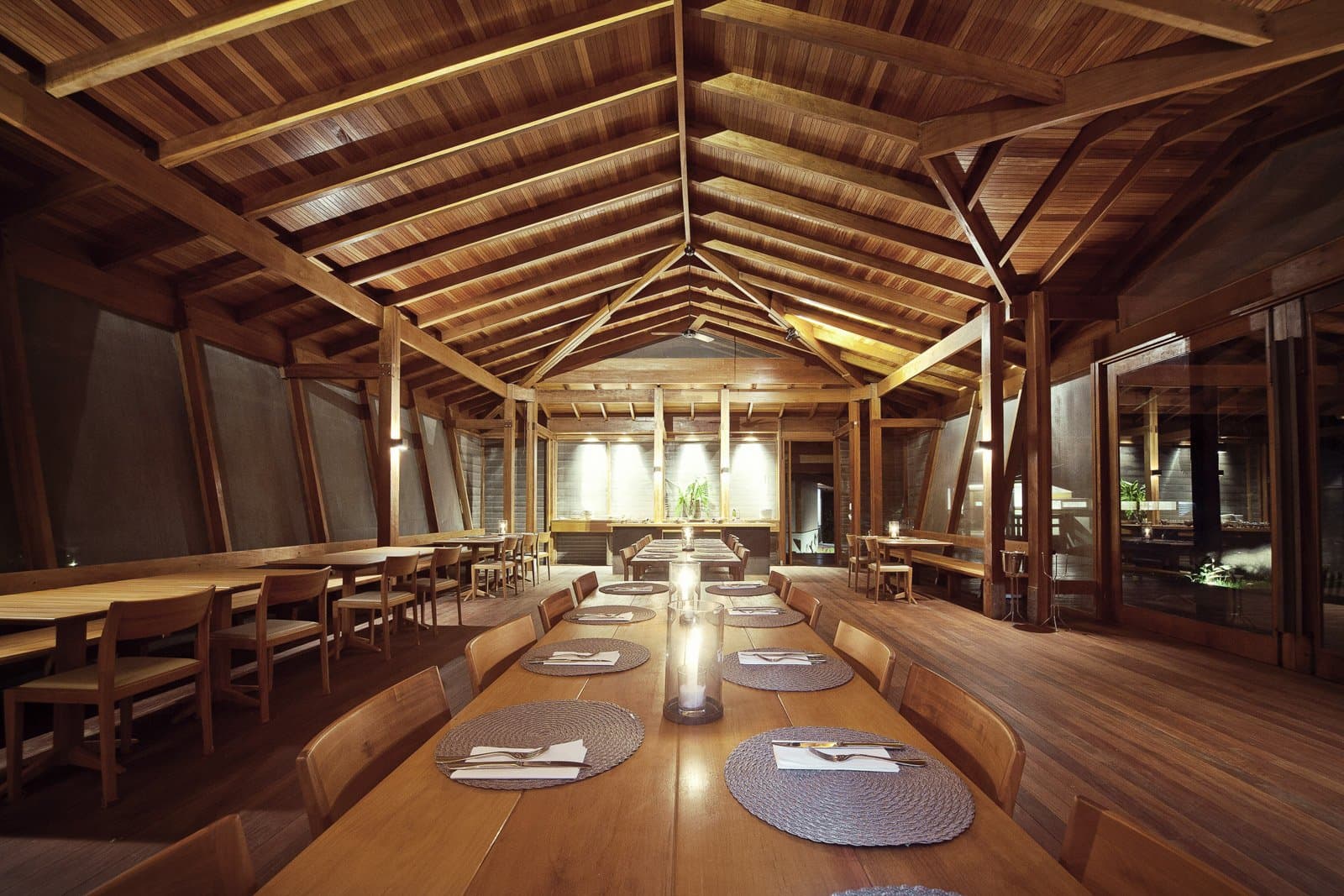 Gutes Essen, gute Gesellschaft, und eine Decke, die für sich spricht. Photo: Samuel Melim (c) Warm beleuchteter Holzspeisesaal mit gewölbter Holzdecke, langer Gemeinschaftstafel mit geflochtenen Tischsets und Kerzen sowie kleineren Tischen an den Seiten