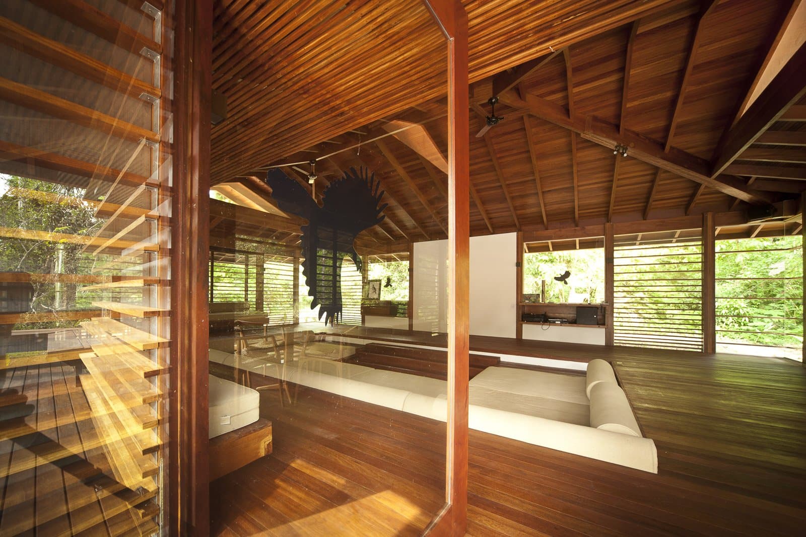 Holz, weiße Kissen, Wald wohin man schaut. Hier lässt man den Tag ruhig ausklingen. Photo: Samuel Melim (c) Offener Gemeinschaftsraum einer Lodge für eine Luxusreise in den Pantanal und Amazonas, mit warmen Hartholzböden, freiliegenden Holzbalken, weißen Liegesofas, Lamellenfenstern auf drei Seiten und einer großen dekorativen Vogelskulptur an der Decke, umgeben von dichtem grünem Regenwald