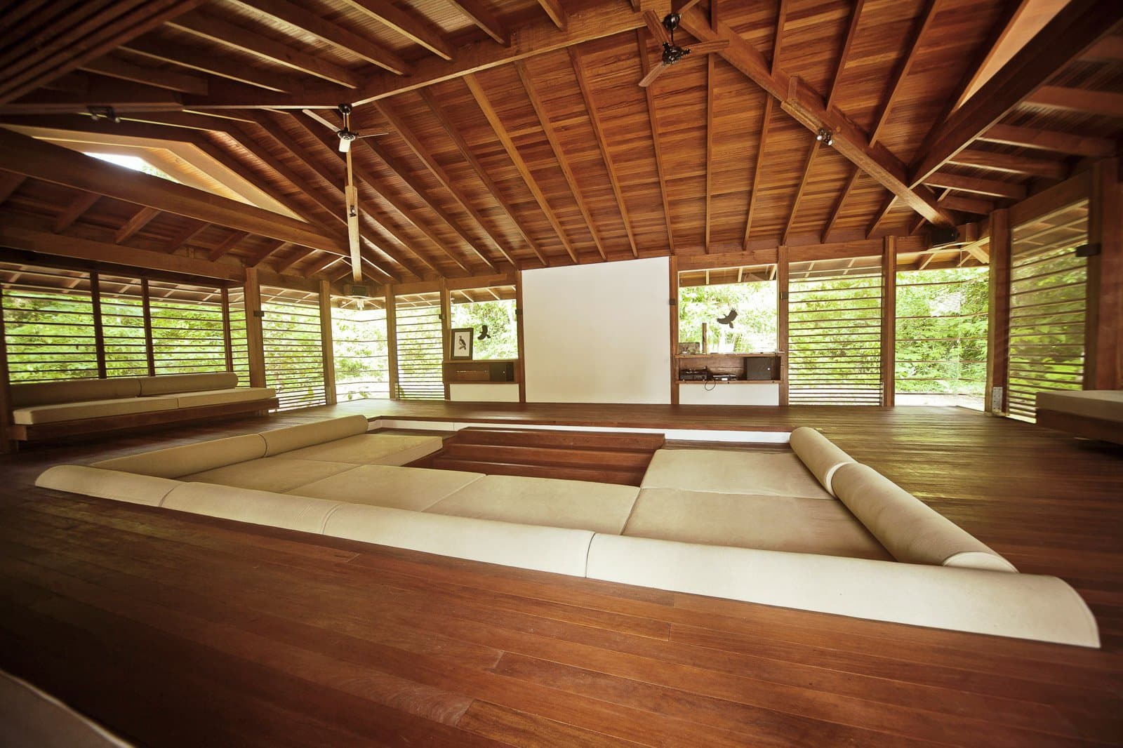 In die Kissen sinken, während der Wald durch die Wände strömt. Photo: Samuel Melim (c) Offene Holz-Lodge-Lounge auf einer Luxusreise durch Pantanal und Amazonas, mit einem versenkten cremefarbenen Ecksofa auf Hartholzboden unter einer hohen Holzdecke mit Deckenventilatoren und vergitterten Wänden zum üppigen Wald hin