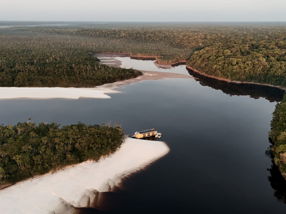Veduta aerea di una piccola imbarcazione fluviale ormeggiata su un banco di sabbia bianca lungo un fiume scuro dell'Amazzonia, circondata dalla foresta.