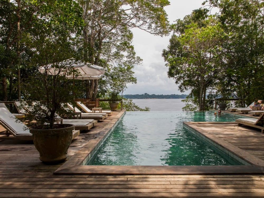 . Uma piscina de borda infinita em um deck de madeira, com espreguiçadeiras, com vista para um rio, emoldurada por árvores da floresta em um ecolodge na Amazônia.
