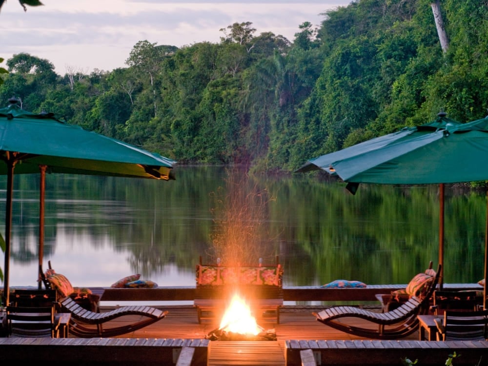 . Um deck de pousada à beira-rio ao anoitecer, com guarda-sóis verdes, espreguiçadeiras e uma fogueira acesa refletida nas águas calmas do rio amazônico.