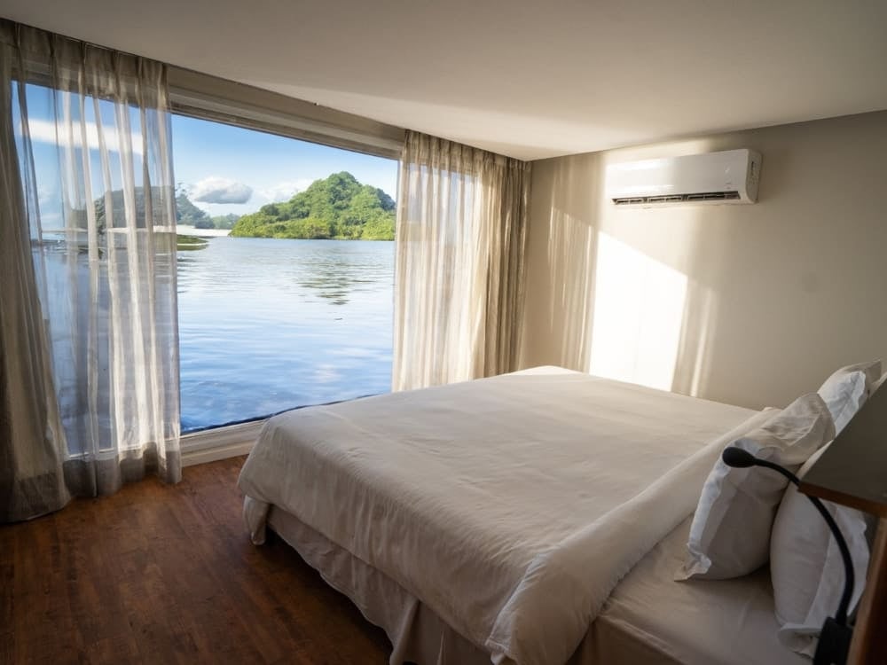 . Um quarto de pousada claro, com cama de casal e janelas do piso ao teto com vista para um rio e uma ilha florestada na Amazônia.