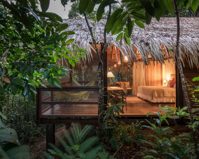 A luxury Amazon jungle lodge bungalow with a thatched roof and glass walls glowing with warm light at dusk, a hammock on its wooden deck amid the rainforest. A luxury Amazon jungle lodge bungalow with a thatched roof and glass walls glowing with warm light at dusk, a hammock on its wooden deck amid the rainforest.