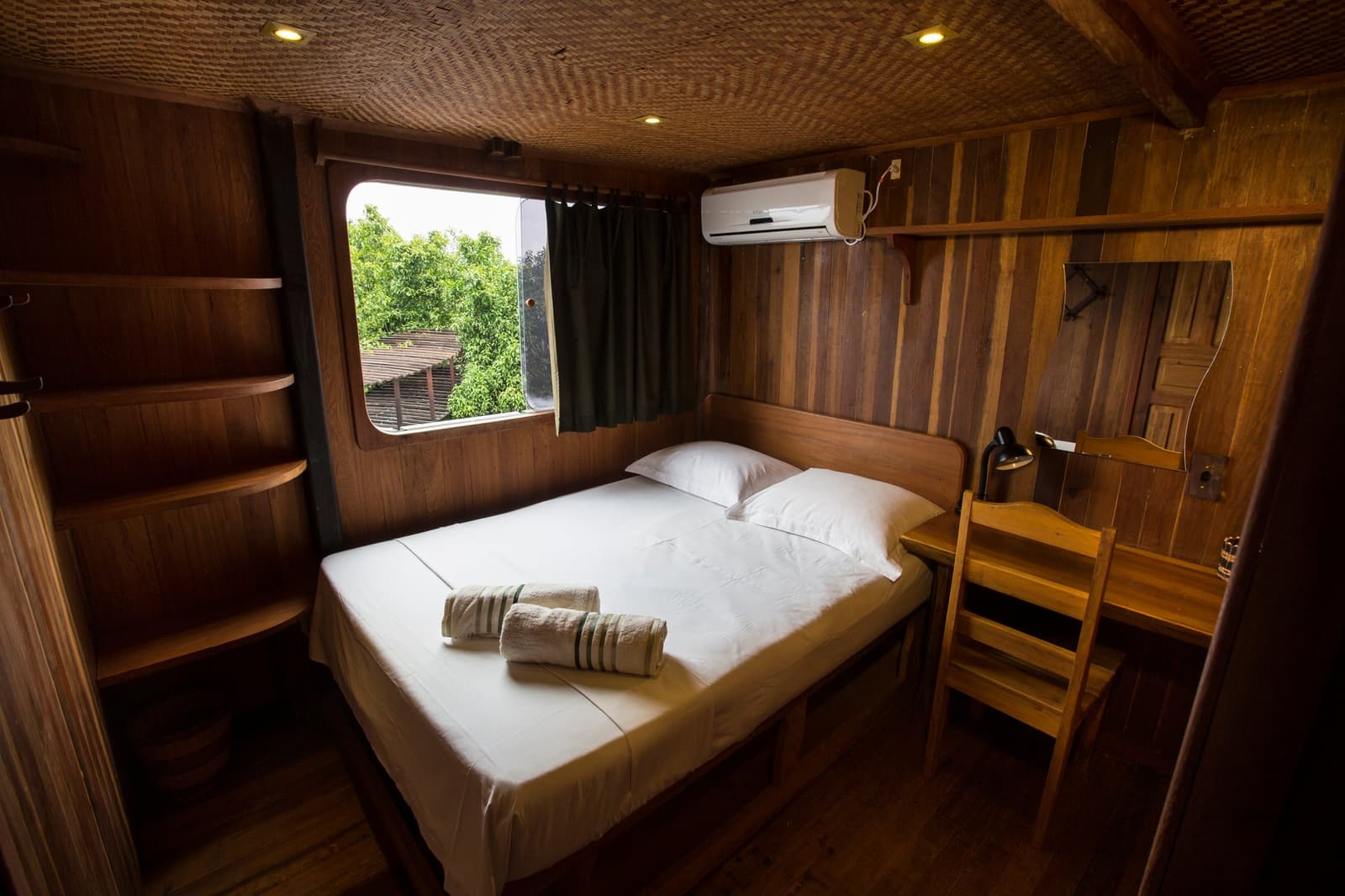 Tudo no lugar certo, e a floresta ali do lado da janela. Cabine aconchegante com paredes de madeira em um cruzeiro pelo Rio Amazonas no Brasil, com cama de casal em roupa de cama branca, toalhas enroladas, prateleiras abertas, escrivaninha, ar-condicionado e janela redonda com vista para a vegetação ribeirinha