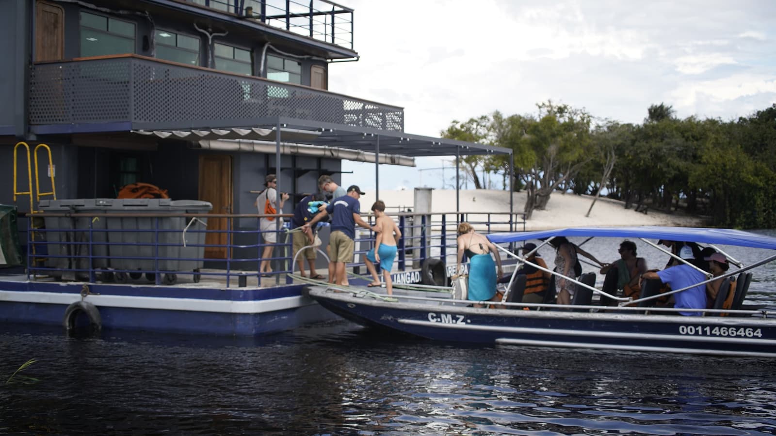 Tutti a bordo, poi ci pensa il Rio Negro. Ospiti che salgono su un motoscafo blu al pontile galleggiante della crociera amazzonica La Jangada, con il catamarano in acciaio a due piani e una riva sabbiosa sullo sfondo