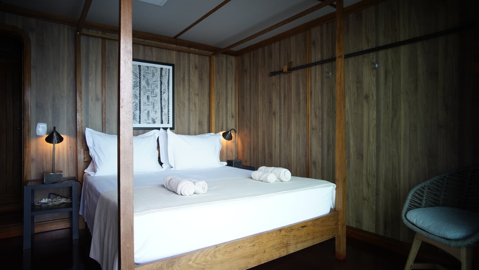 Crisp sheets, warm wood, and nowhere to be until the river calls. Four-poster wooden bed with white linens and rolled towels on a la jangada amazon cruise cabin, warm bedside lamps and indigenous-style artwork on wood-panelled walls
