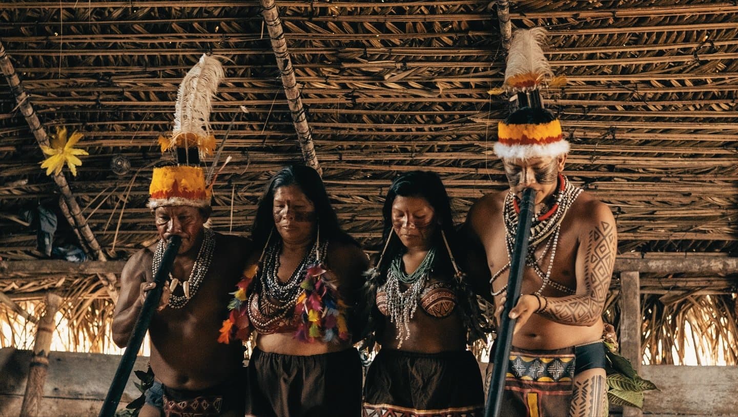 Secoli di sapere, indossati con orgoglio silenzioso. Quattro membri di una comunità indigena con copricapi di piume, collane di perle e decorazioni corporee geometriche eseguono una cerimonia all'interno di una maloca dal tetto di paglia