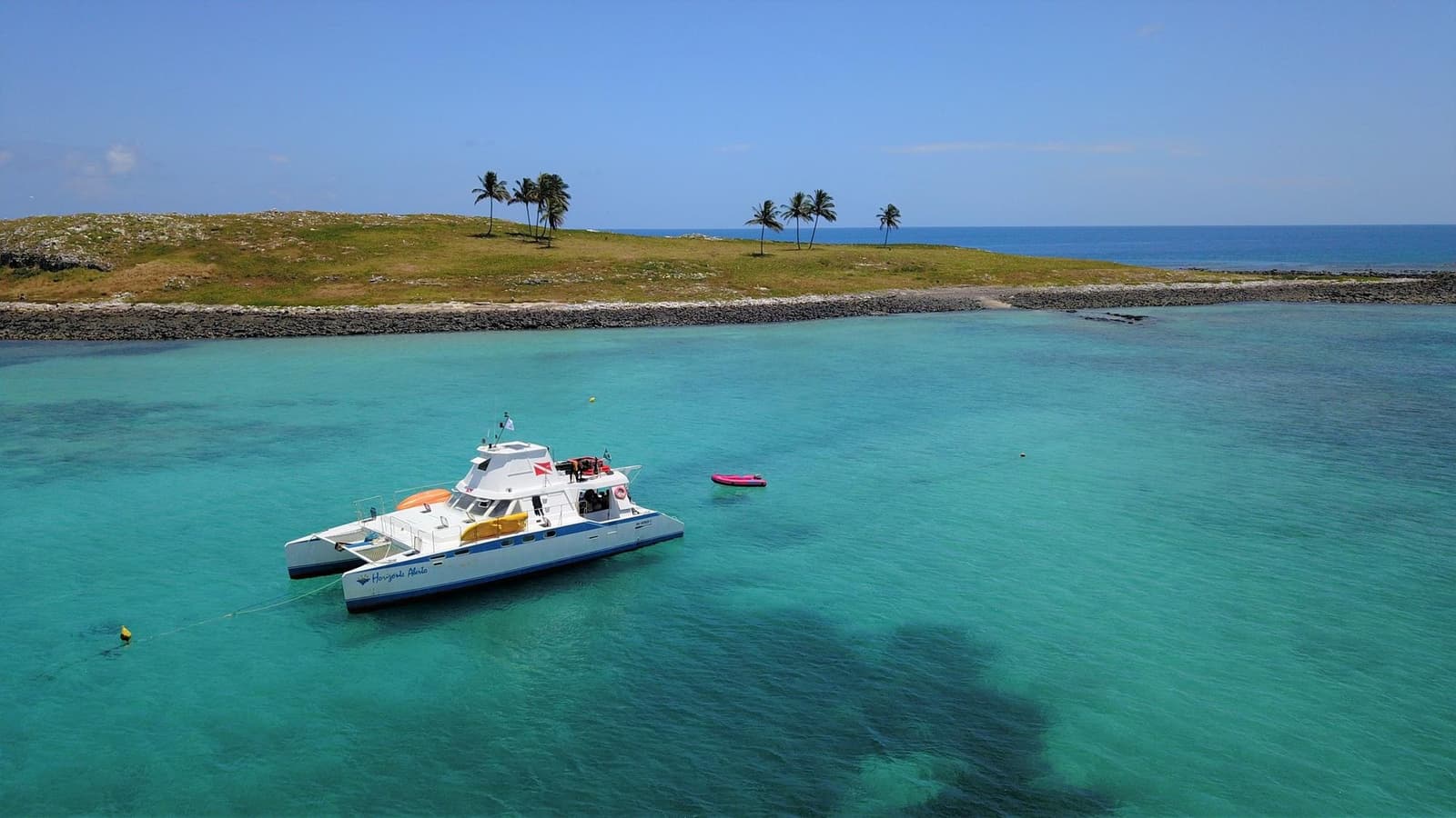 Your home for three days, and the ocean is the backyard. A white catamaran named Horizonte Aberto anchored in a liveaboard Abrolhos setting, floating over turquoise shallow water beside a low palm-lined island under a clear blue sky.