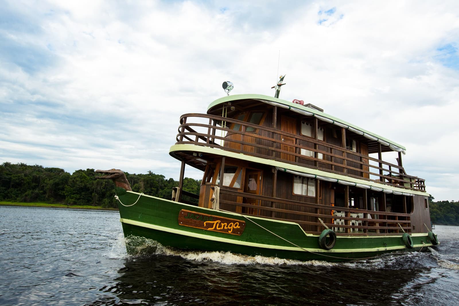 Com o jacaré na proa e o rio na frente, a Tinga segue sem pressa e sem dúvida. Photo: sitah O barco Jacaré-Tinga, com casco verde e três conveses de madeira, navega por um rio de águas negras da Amazônia brasileira à beira de floresta densa