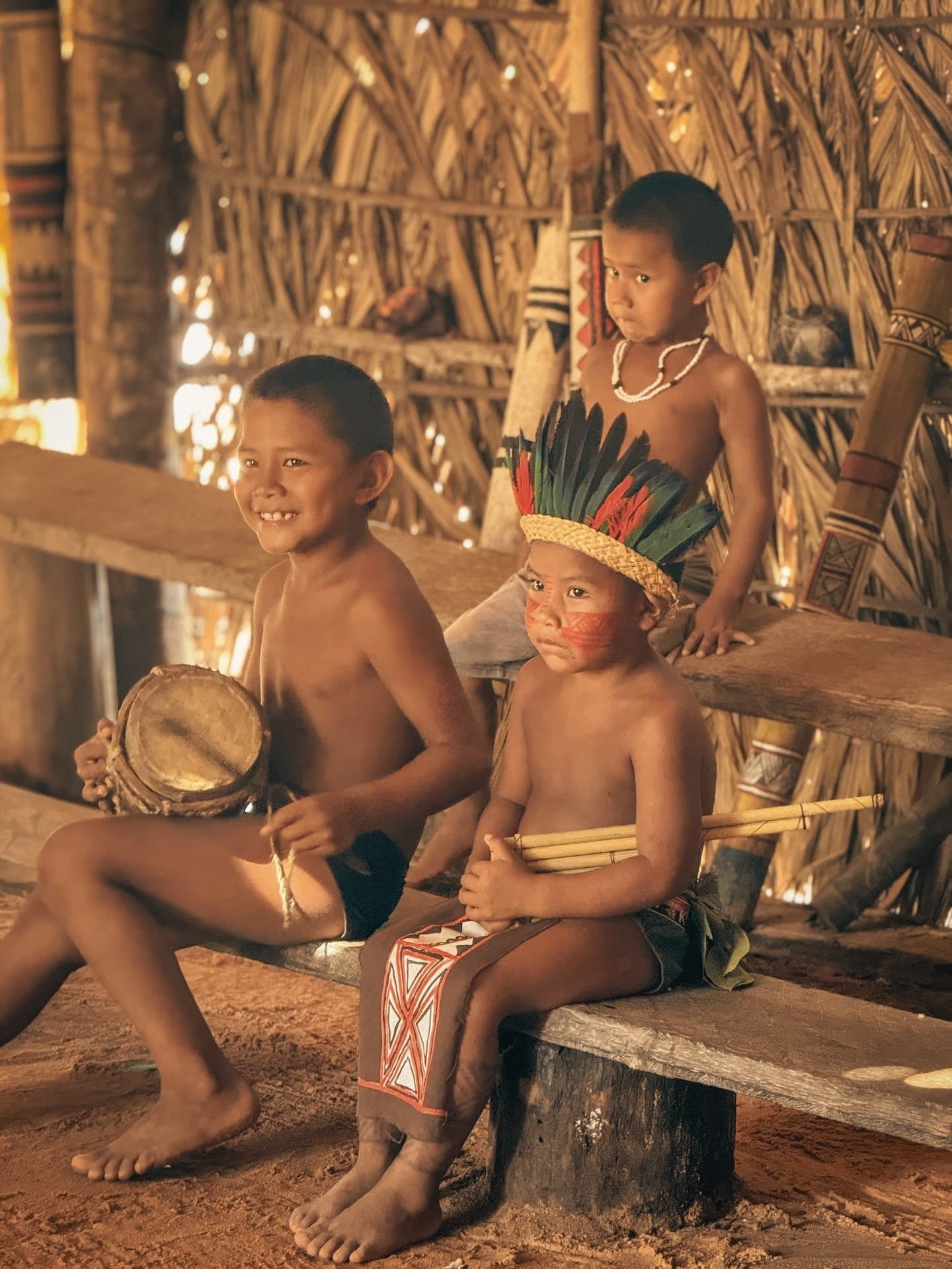 Ogni ritmo, ogni piuma, ogni parola: qui si imparano davvero. Tre bambini indigeni all'interno di una maloca di paglia durante una visita comunitaria della crociera La Jangada sull'Amazzonia — un ragazzo sorridente tiene un piccolo tamburo, un altro indossa un copricapo di piume e una pittura facciale rossa, un terzo osserva in silenzio sullo sfondo